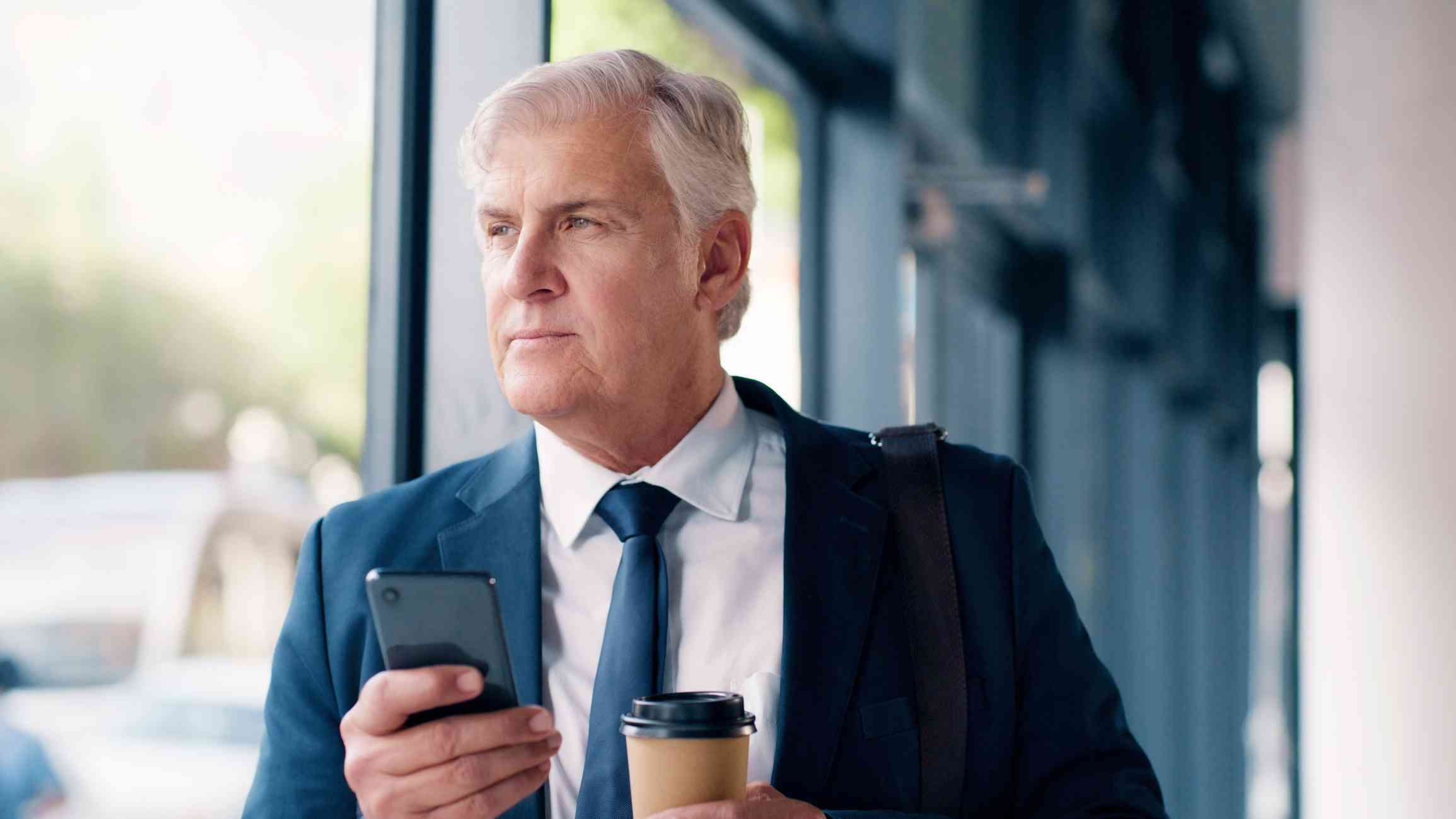 A middle aged man in a buisness suit stands near a window in an office buidling and gazes off deep in thought while holding his phone and a cup of to-go coffee.