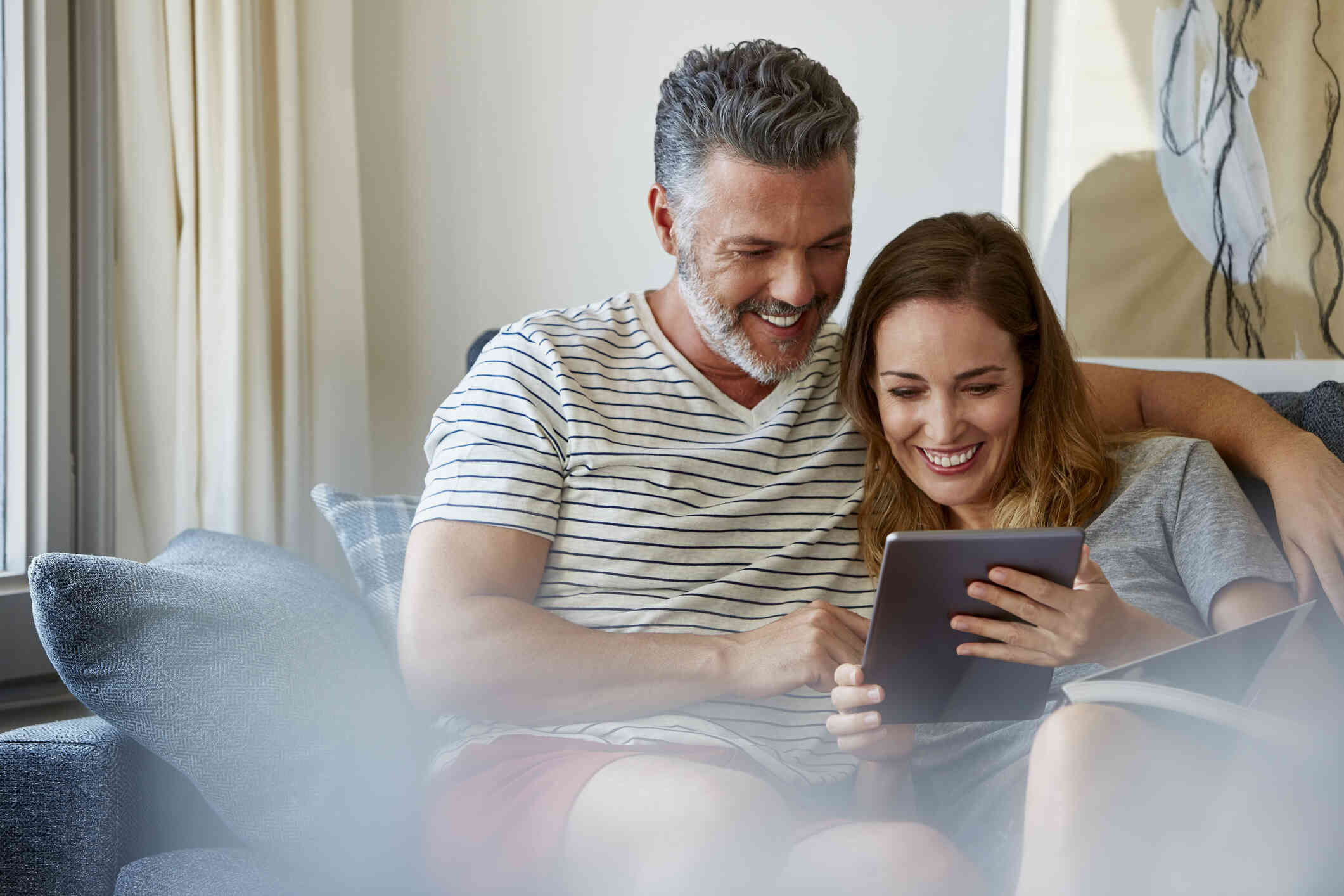 A male and female couple sit close together on the couch while smiling and looking at a tablet together.