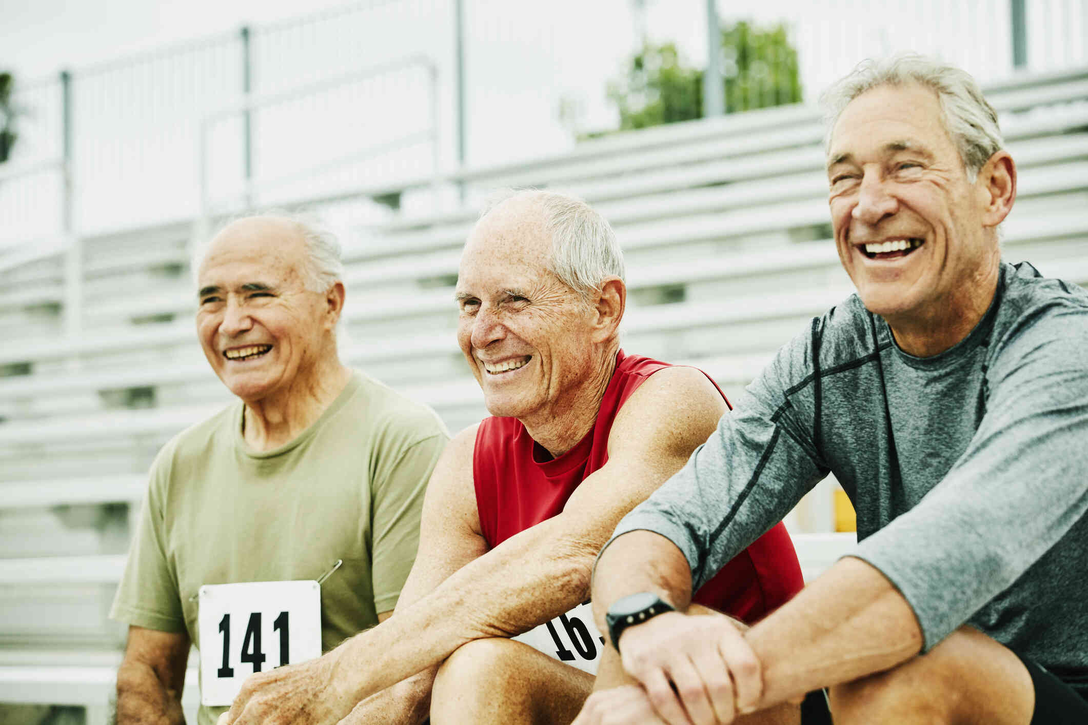 Three elderly men smile while setting next to each other on outdoor bleachers with racing numbers pinned to their shirts.