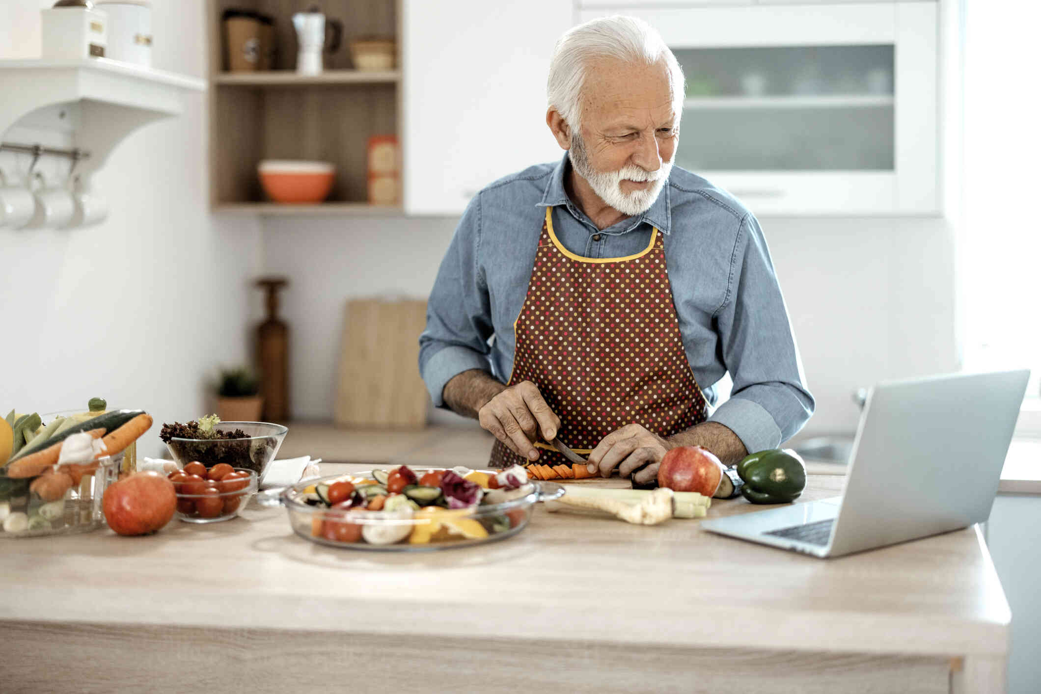 A mature man looks focused as he stands in a kitchen wearing an apron while cutting fruits and vegetables and looking at a laptop screen in front of him.