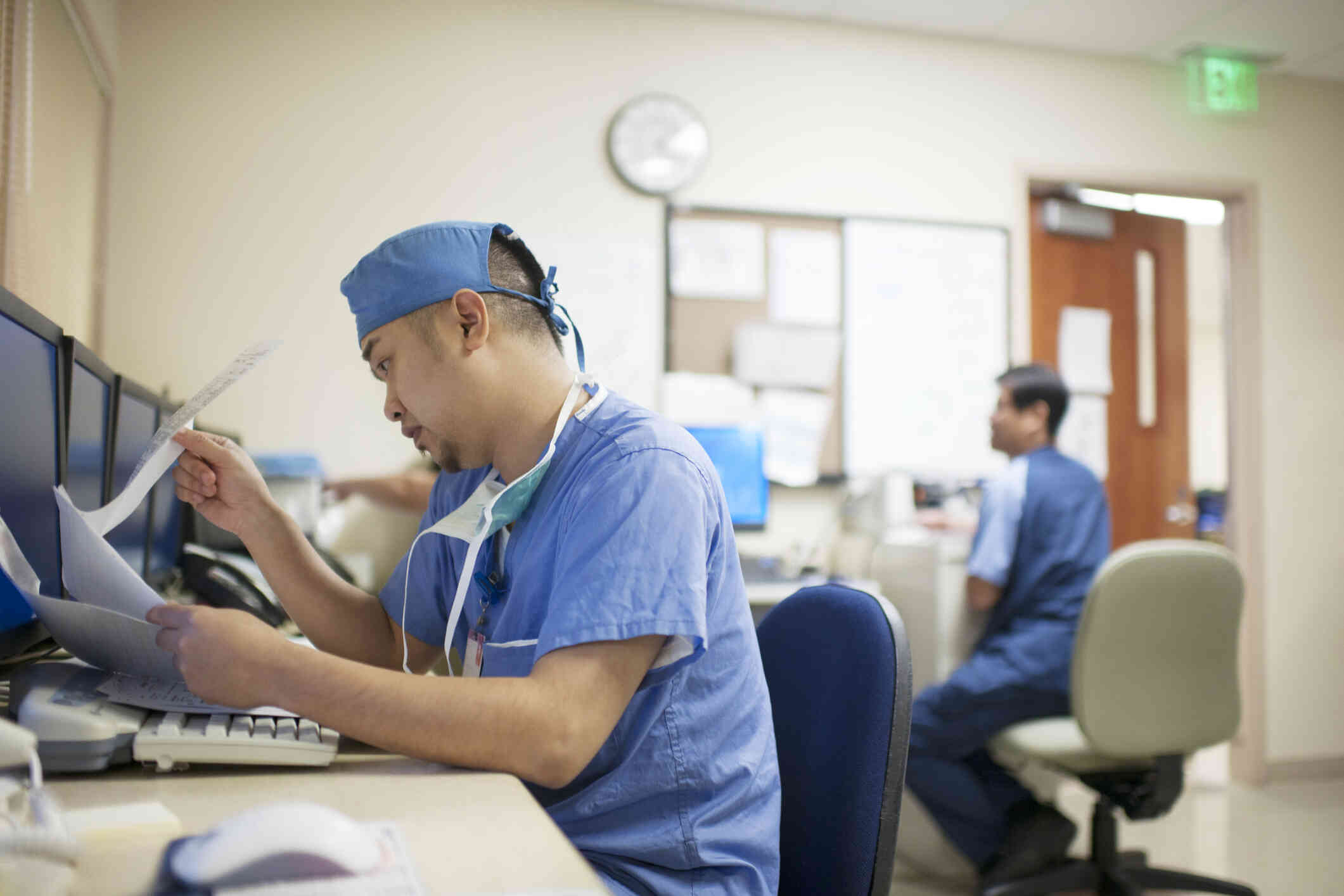 A man in blue, surgery scrubs sits at a desk in an office and looks through paperwork in front of him.