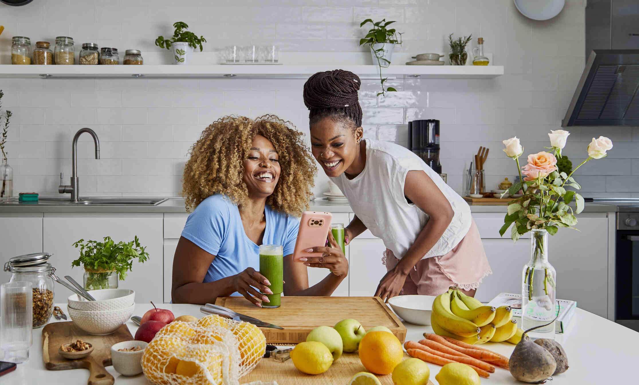 Two women in a kitchen laughing together while looking at a smartphone, surrounded by fresh fruits, vegetables, and a green smoothie.