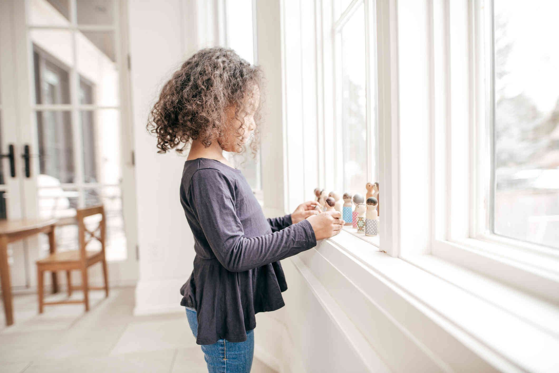 A young girl in a black shirt stands at the window and plays with small figurings in the windowsill.