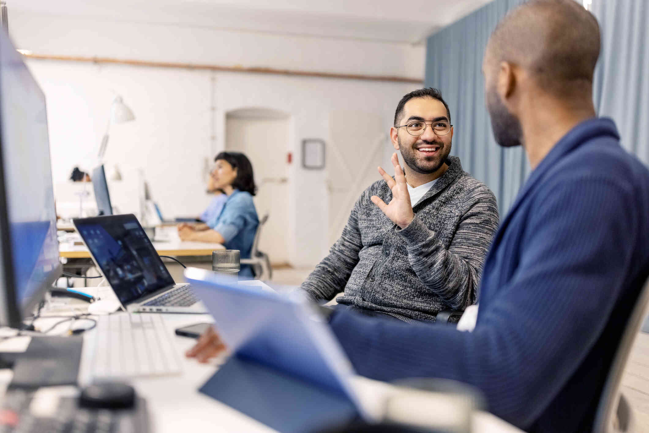 A man in a grey  sweater sits at his work desk and chats with his male boss sitting next to him.