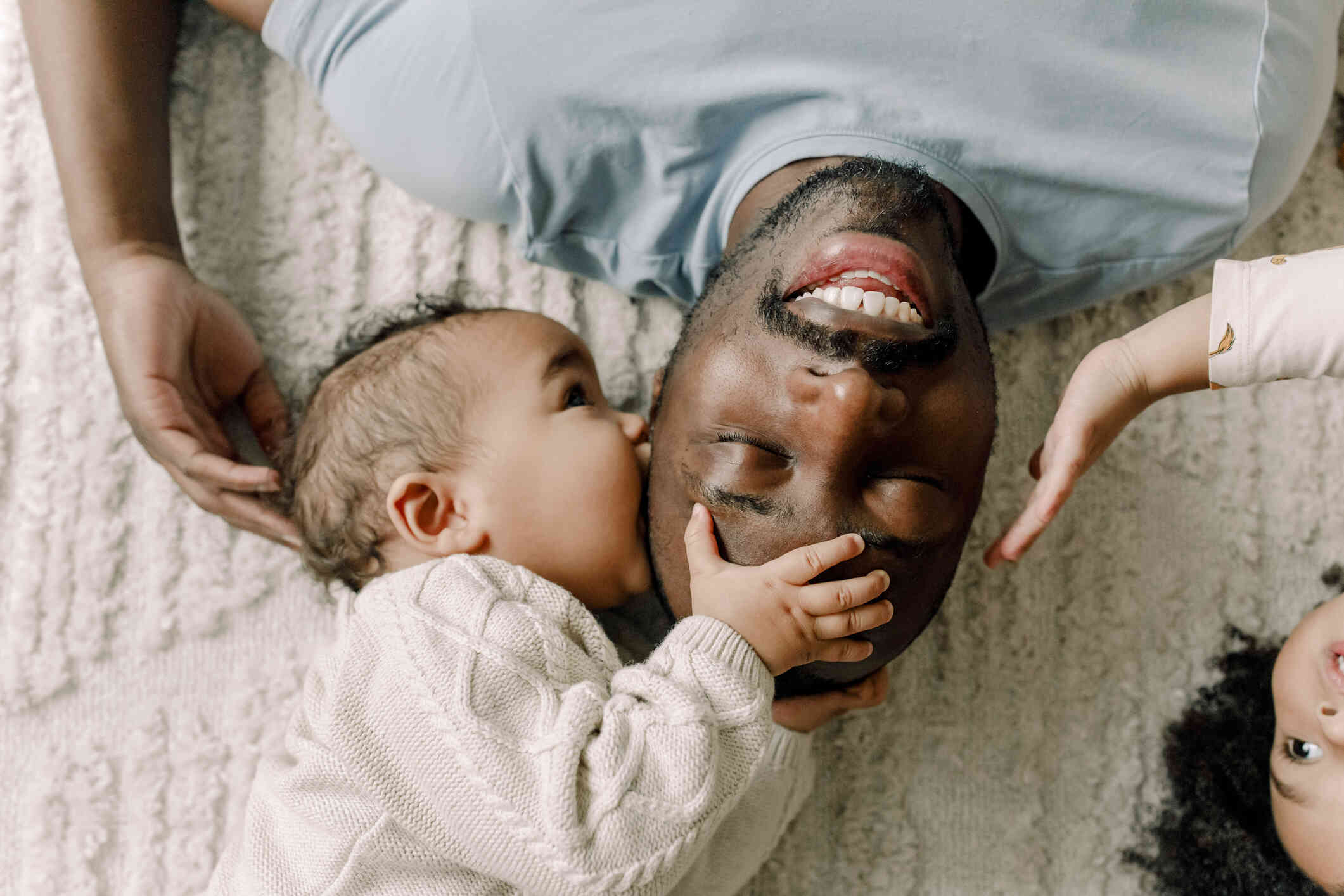 A dad lays on his back on the floor while smilingas his newborn lays next to him while kissing his head.