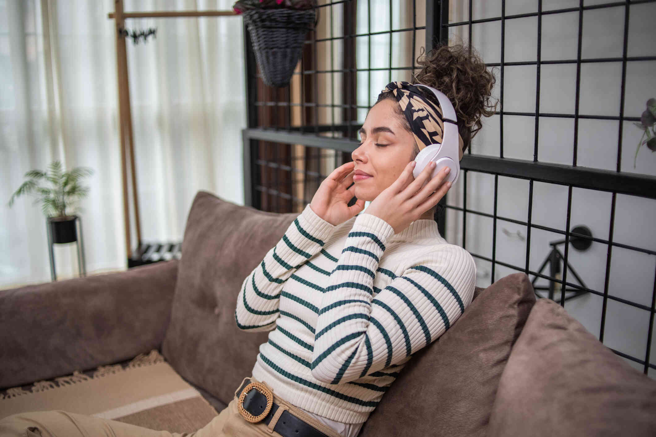 A woman in a striped sweater sits on the couch with white headphones while closing her eyes and gently placing her hands to her ears.