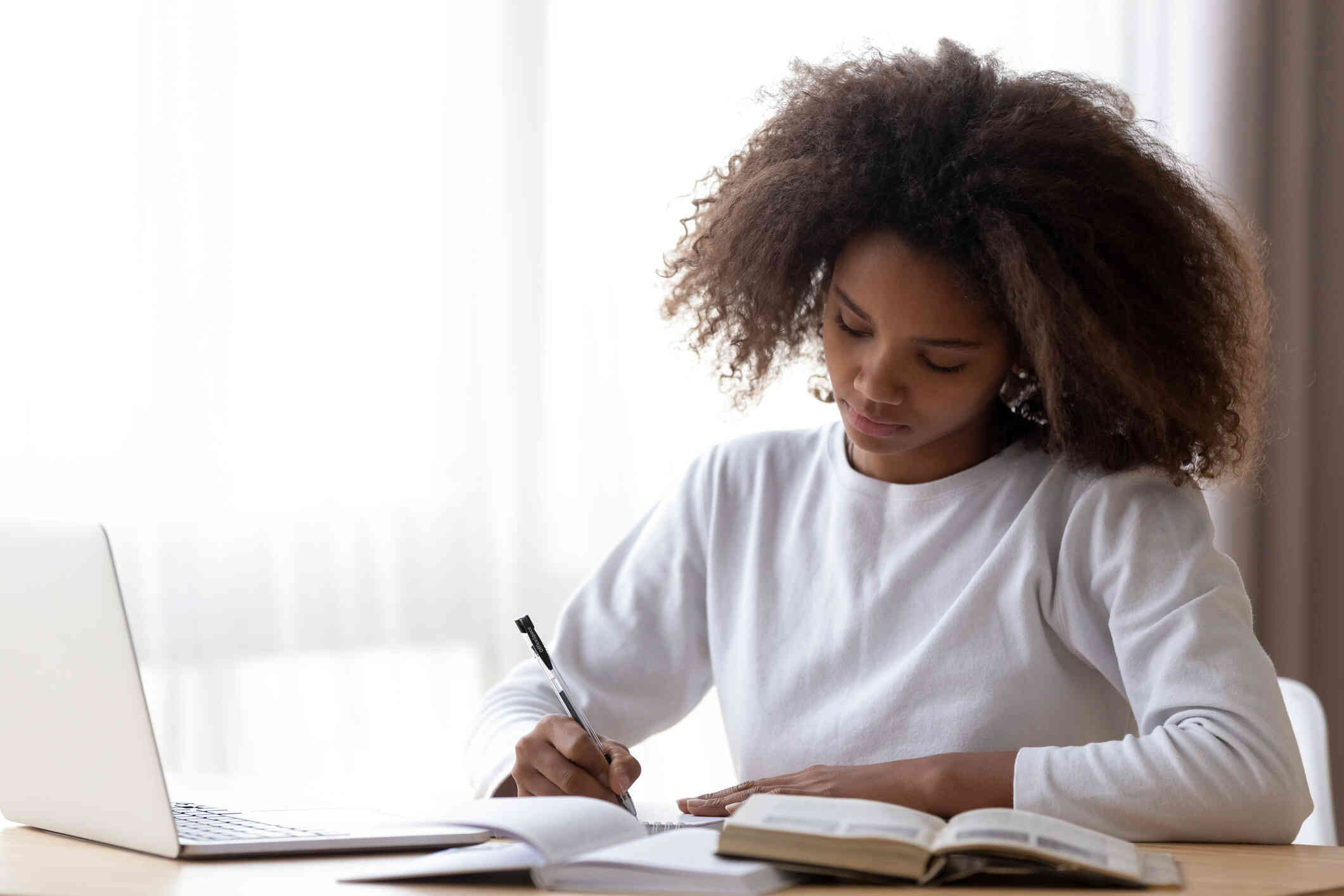 A woman in a white sweater sits at the kitchen table with her laptop and books open next to her as she writes in a notebook.