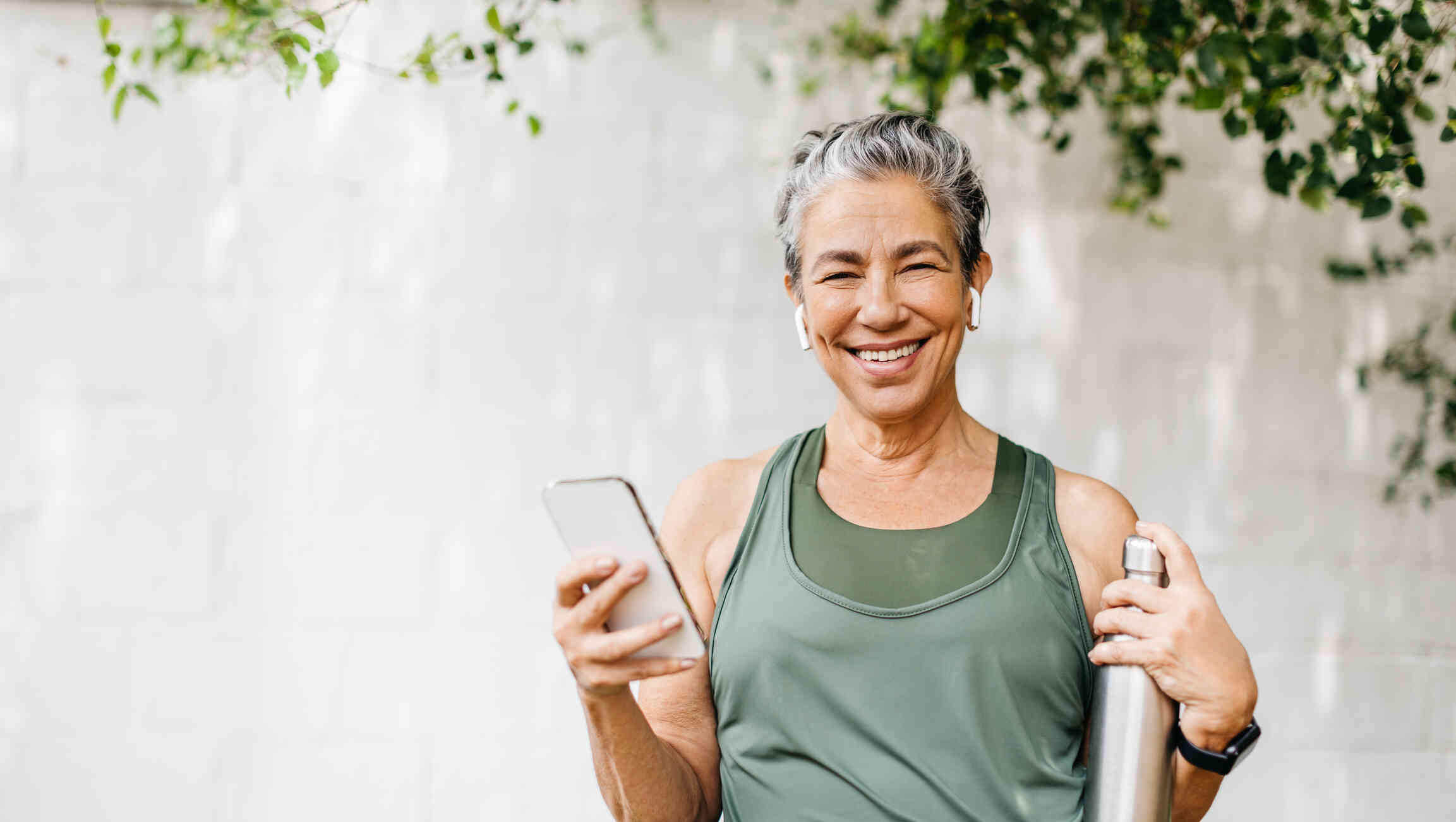 A woman in agreen workout top stands with her phone in her hand and a woterbottle in the other while smiling at the camera.