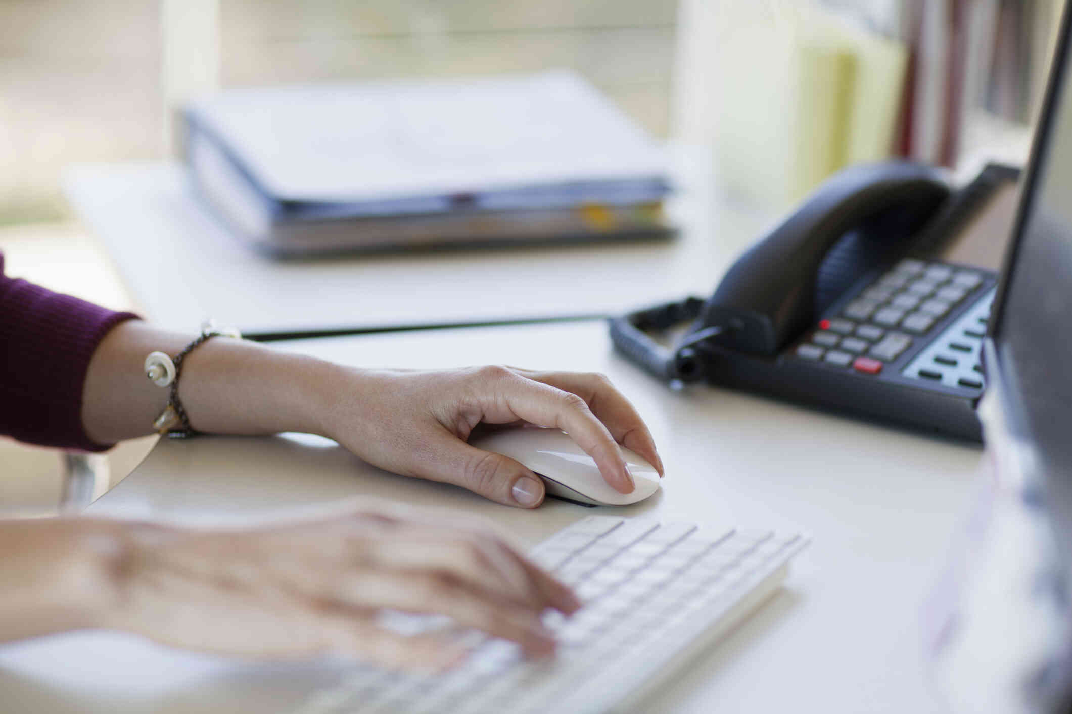 A young woman wearing a maroon shirt types on the computer while also holding the mouse in one hand