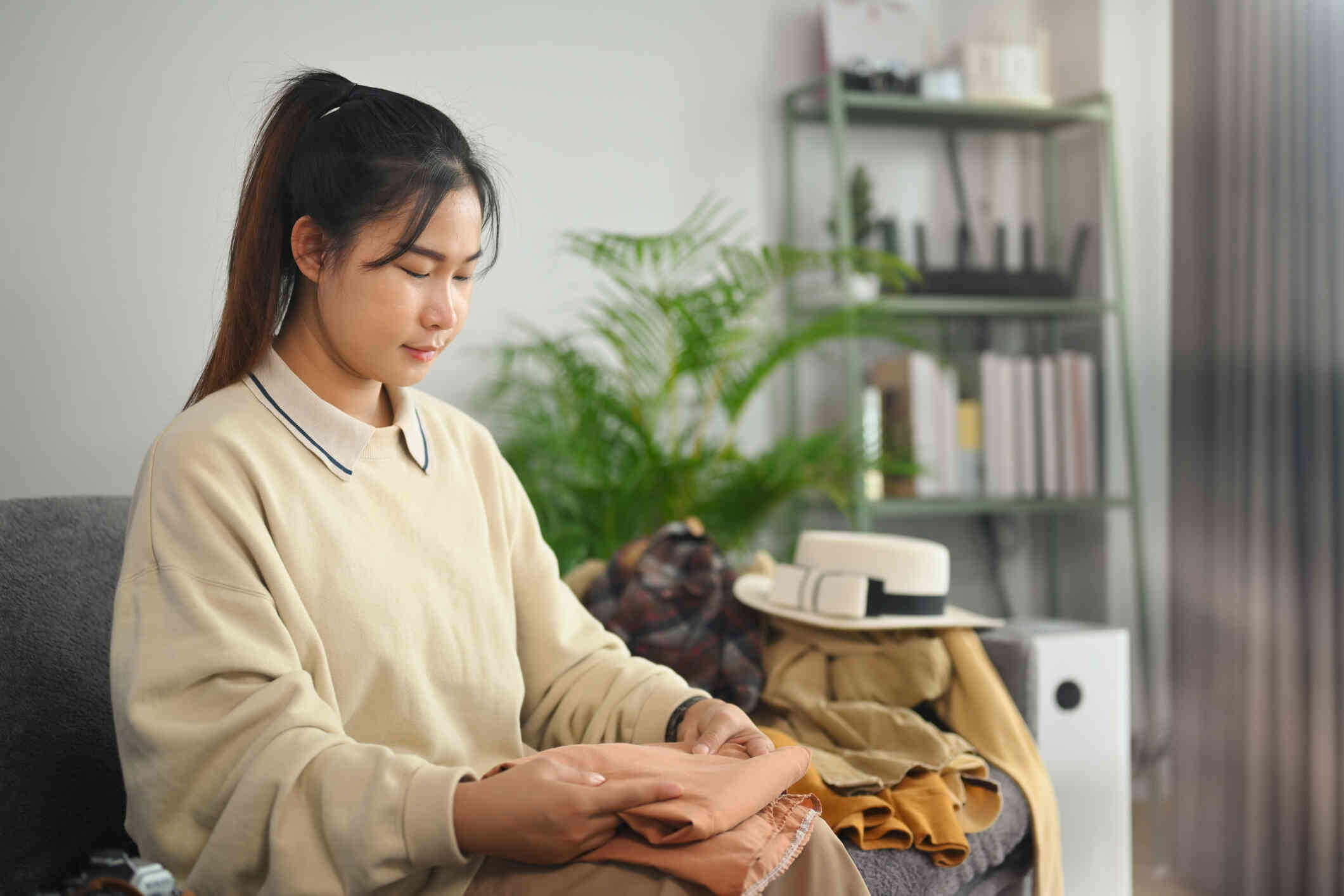 A woman in a tan sweater sits on the couch and folds some clothes in her lap.