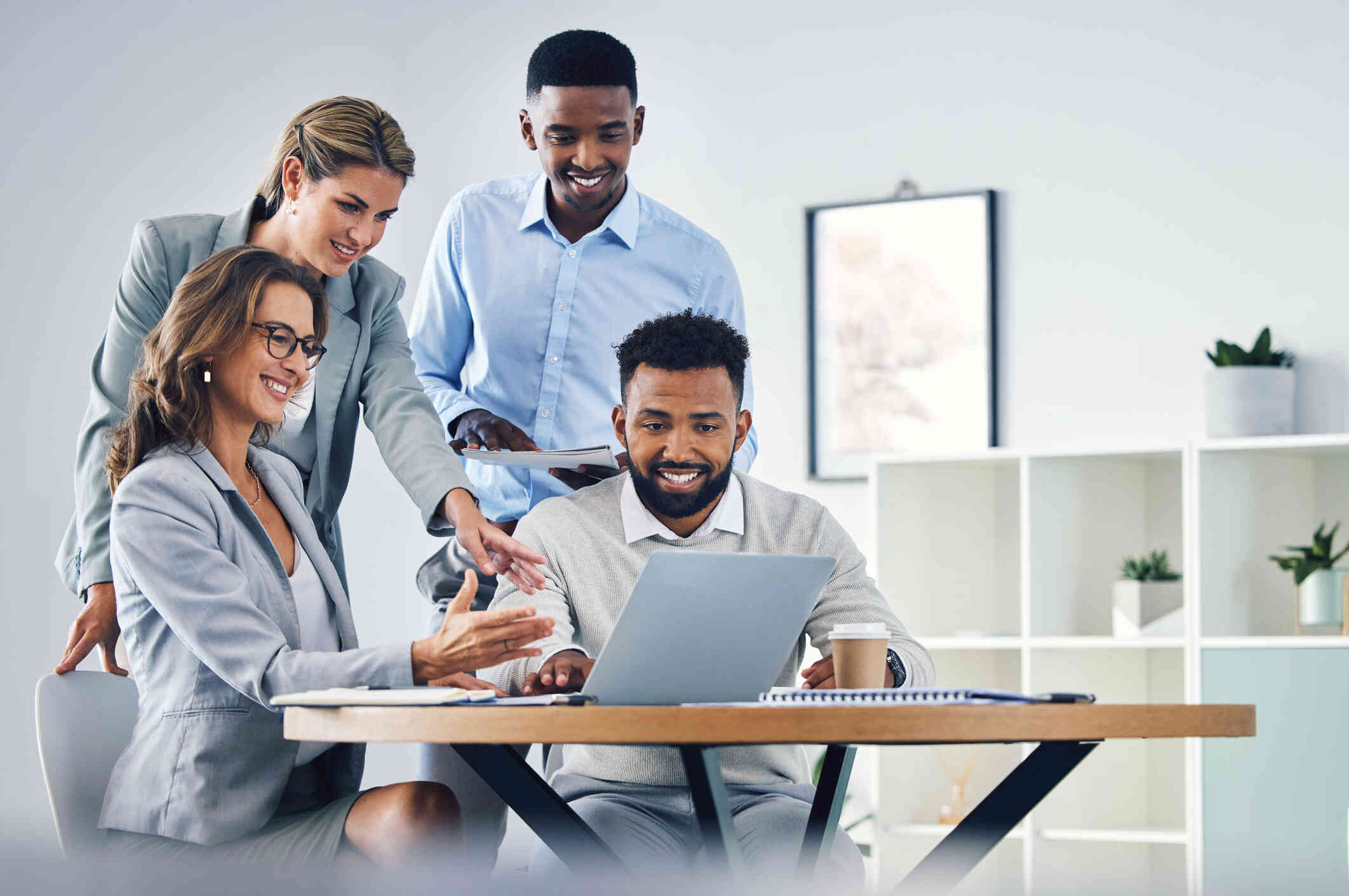 A group of colleagues in formal attire smile as they look at a laptop in an office setting.