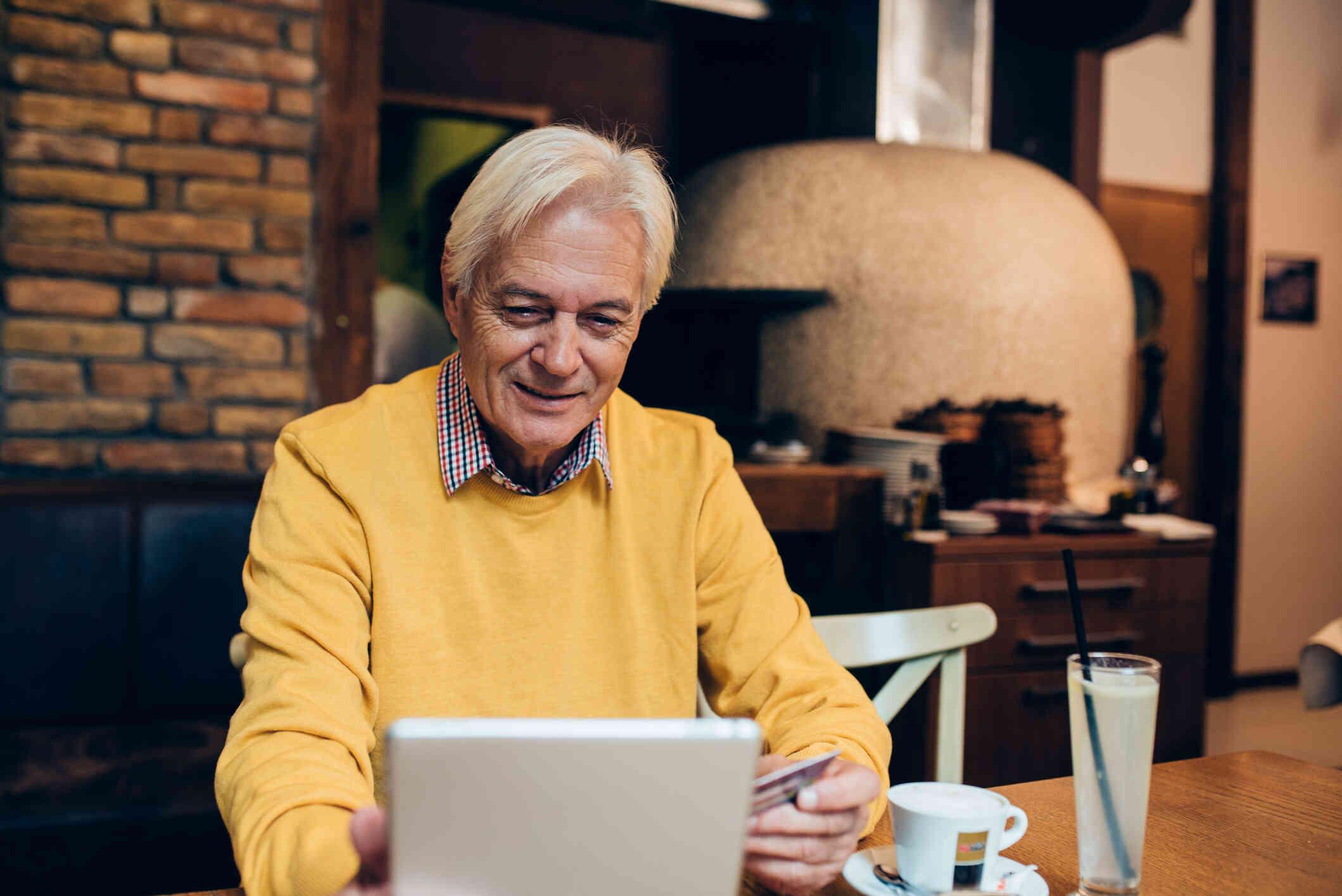 An older man in a yellow sweater, seated at a table in a café, using a tablet with a thoughtful expression.