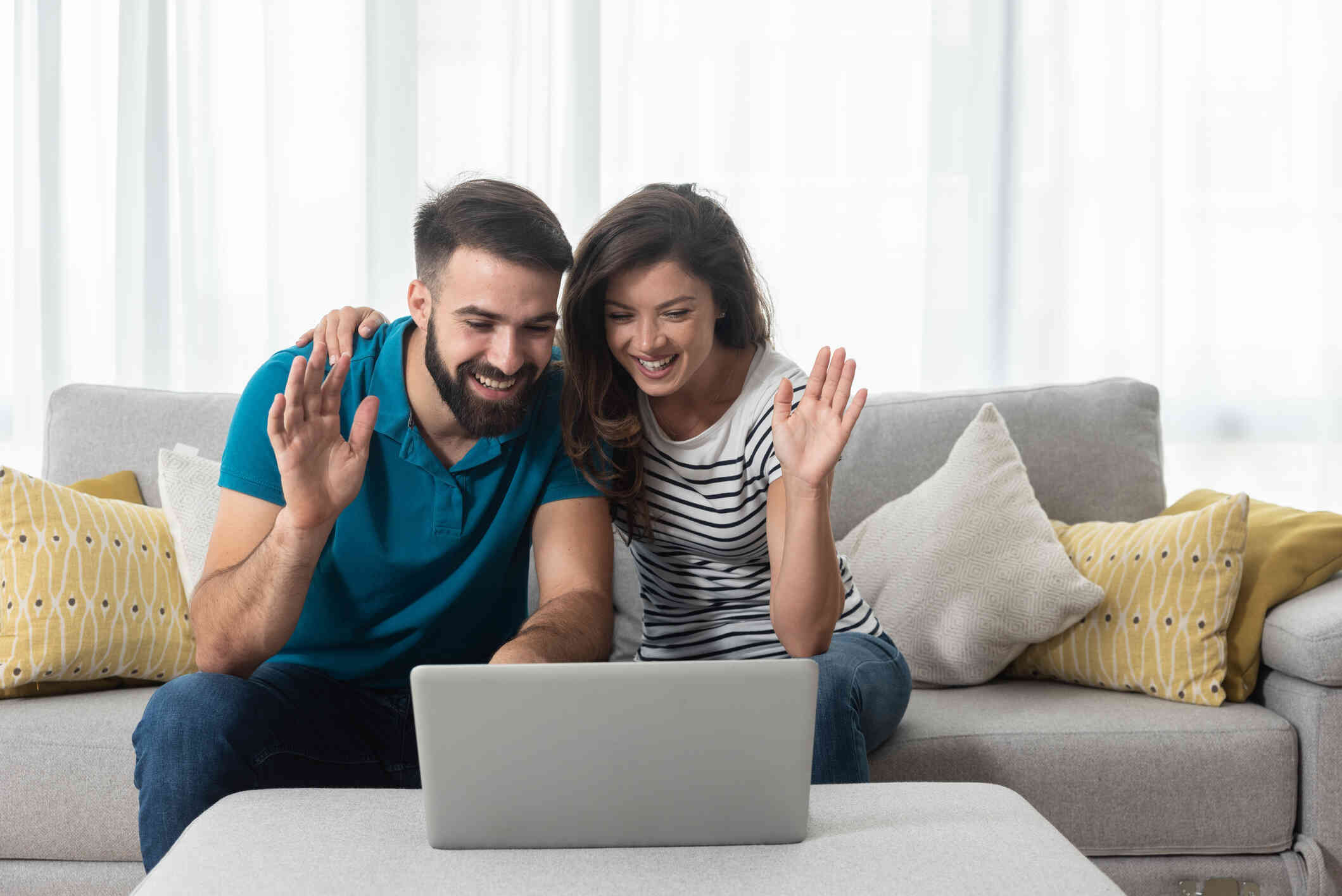 A couple seated on a couch smiles and waves during a video call on their laptop.