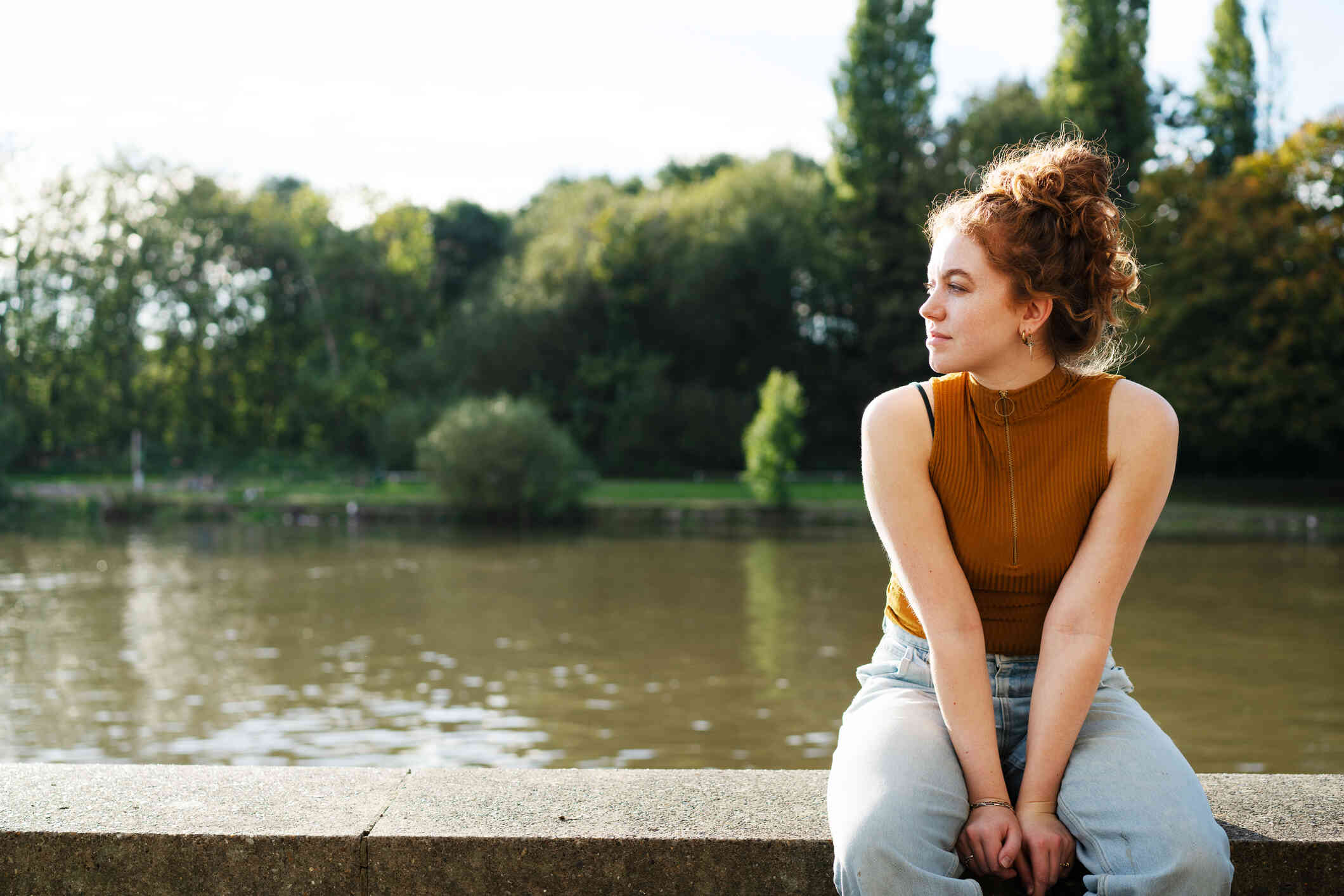 A woman in an orange top sits on a cement ledge near a body of water and gazes off while deep in thought.