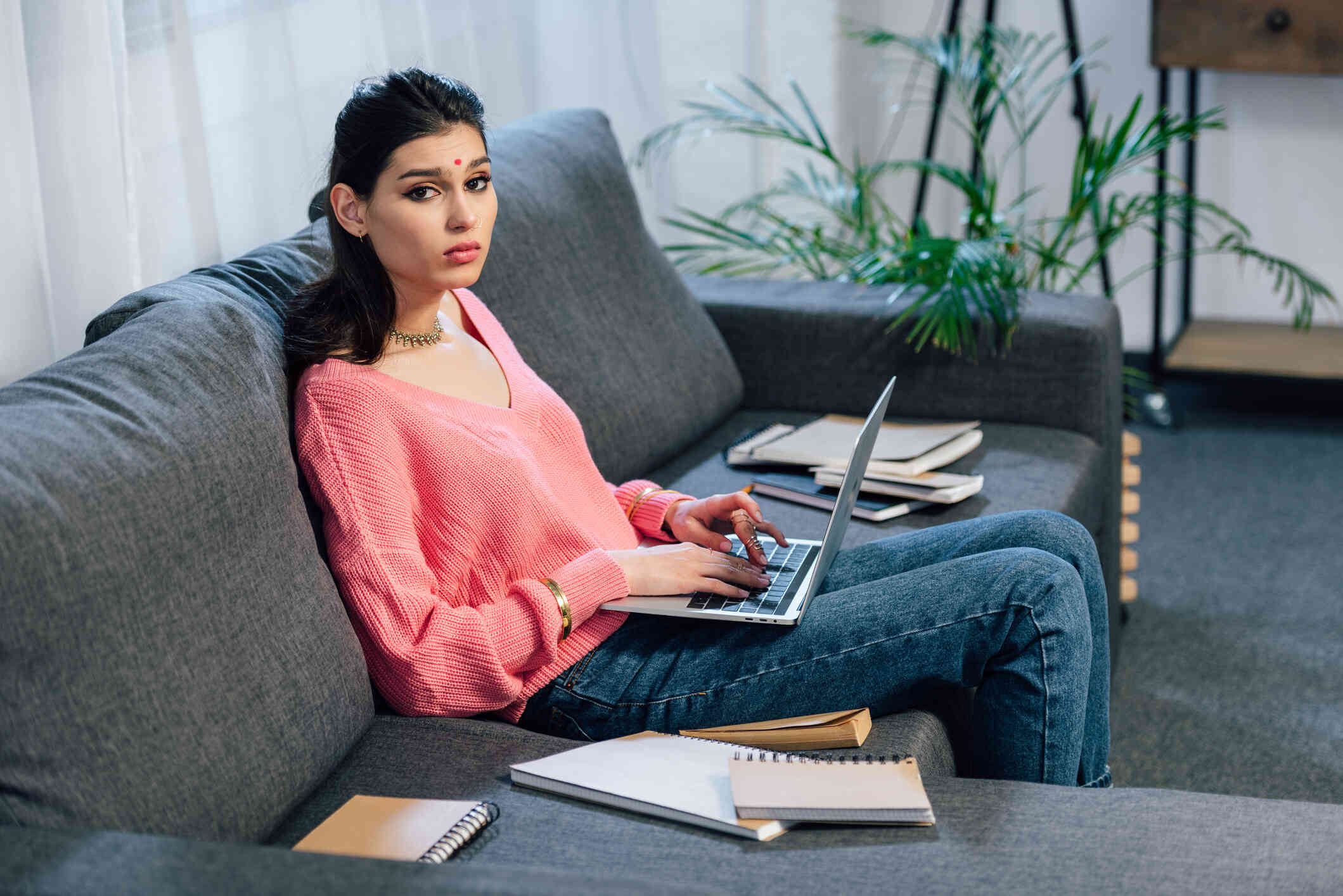 A woman in a pink shirt sits on a grey couch and types on the laptop open in her lap while looking at the camera.