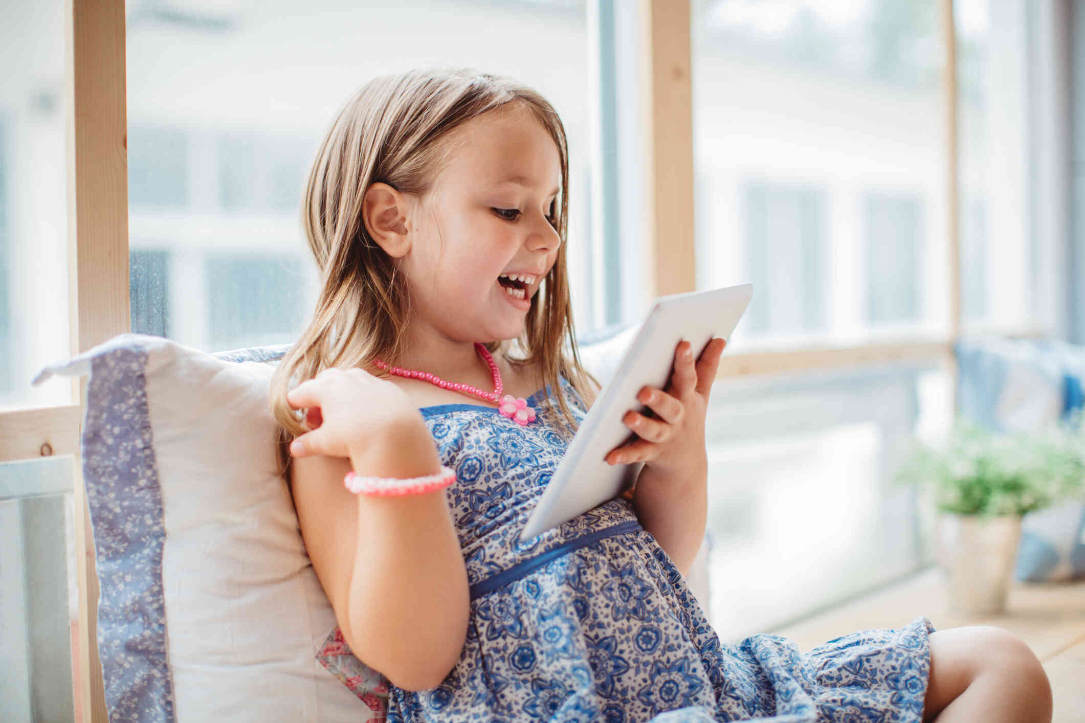 A young girl with blonde hair wears a dress and smiles as she sits on a window seat and looks down at a tablet she is holding.