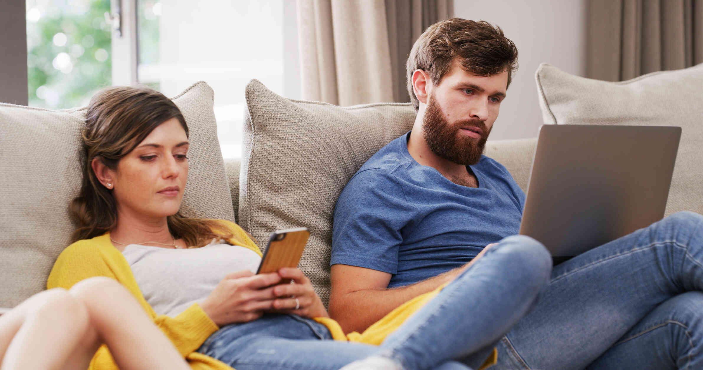 A woman in a yellow cardigan looks down at her phone with a serious expression as she rests on the couch. A man on a laptop sits next to her on the couch.