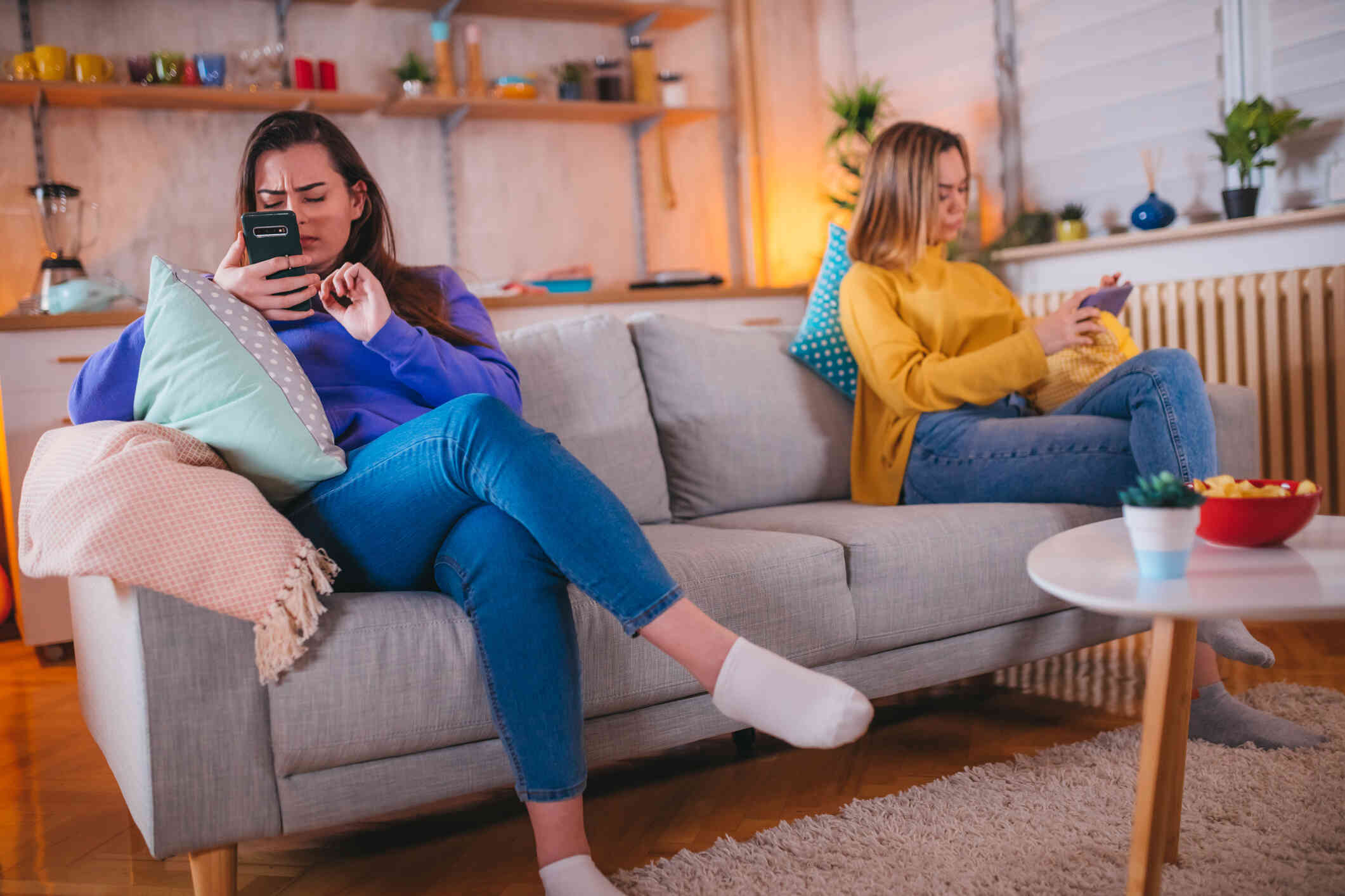 A female couple sit on opposite side of the couch while looking at their own phones and ignoring each other.