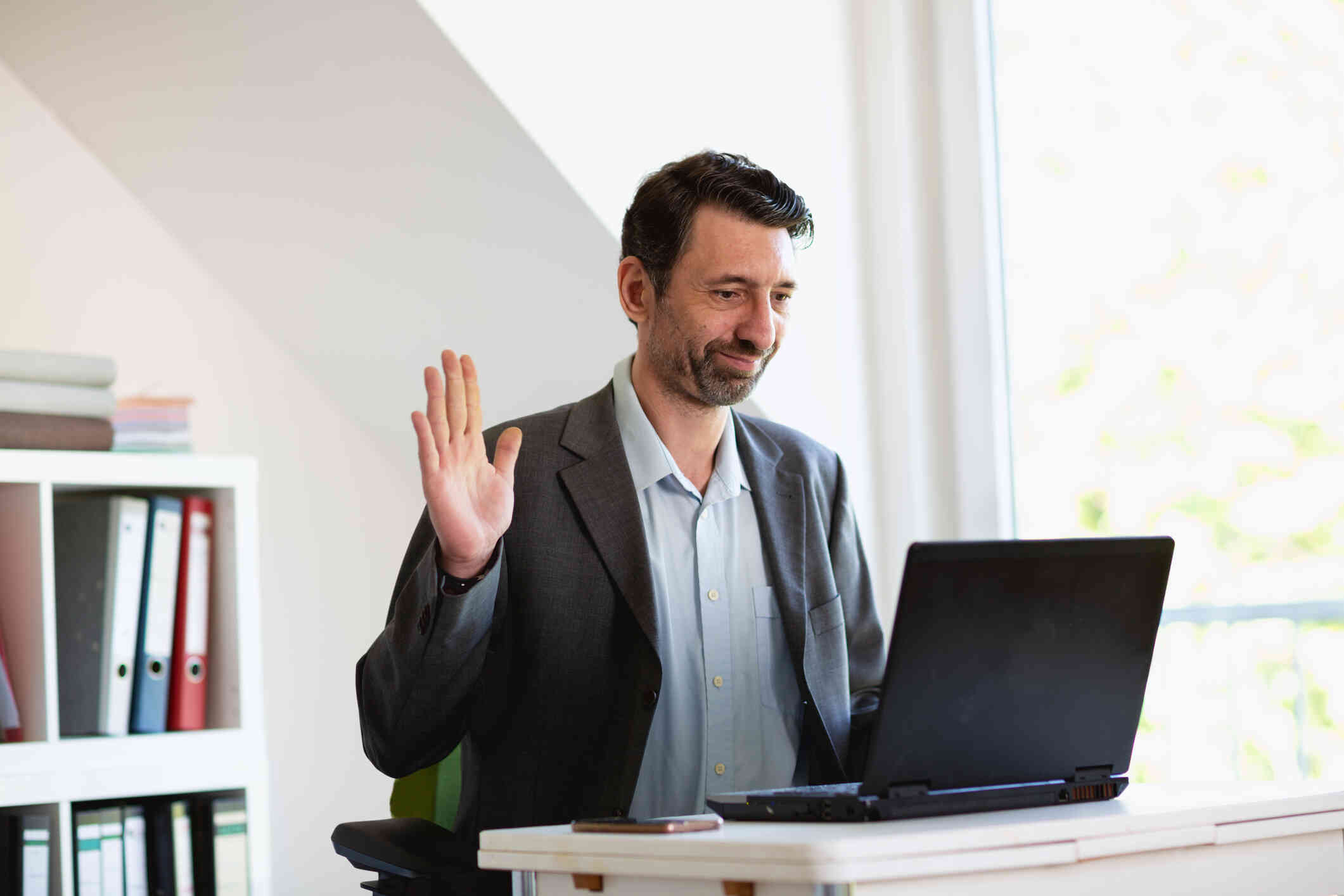 A man in a suit jacket sits at a desk and smiles as he waves his hand towards a laptop which is sitting open in front of him.