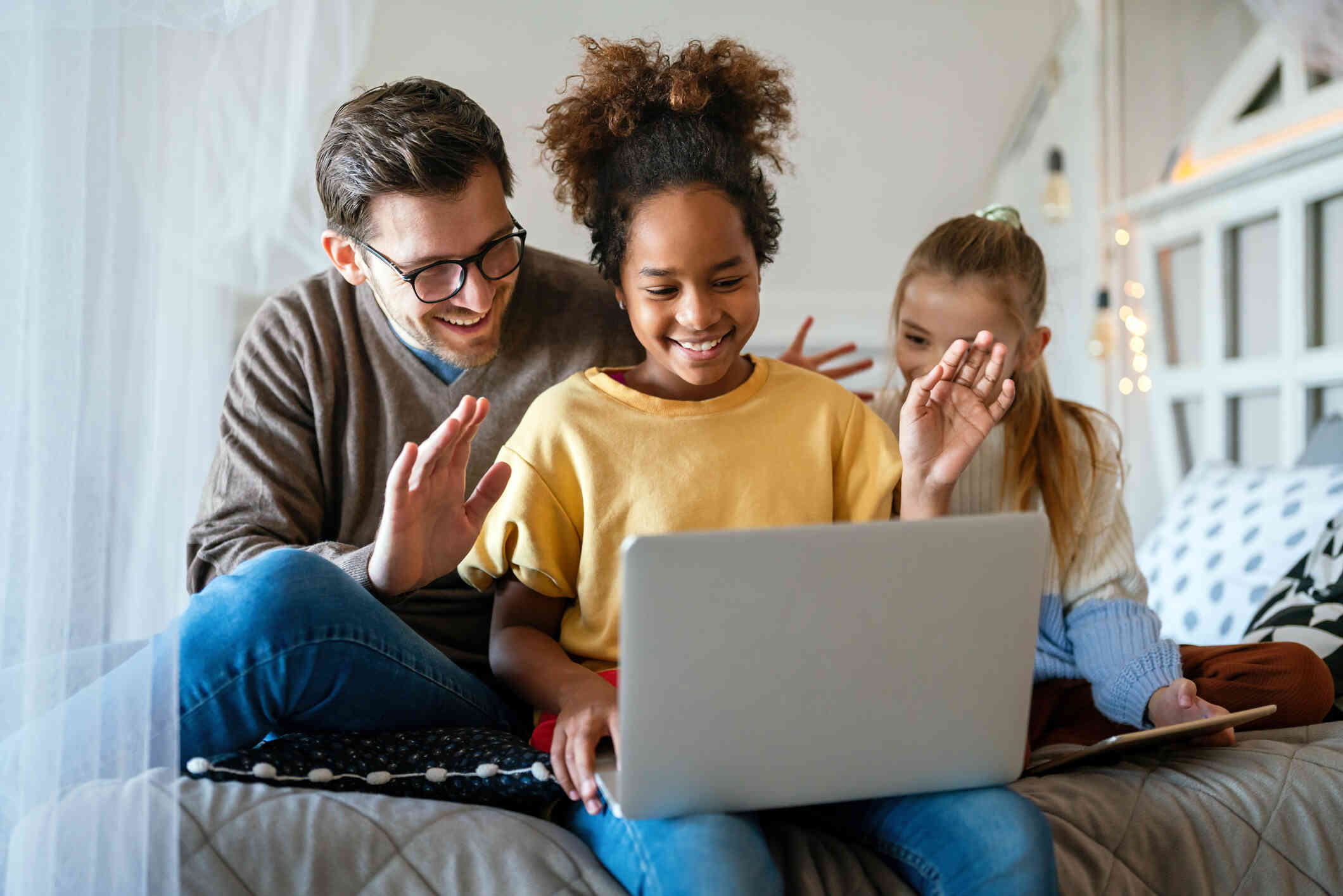 A father and his two daughters sit on a bed and wave at the lpatop open in the girls lap while smiling during a video call.