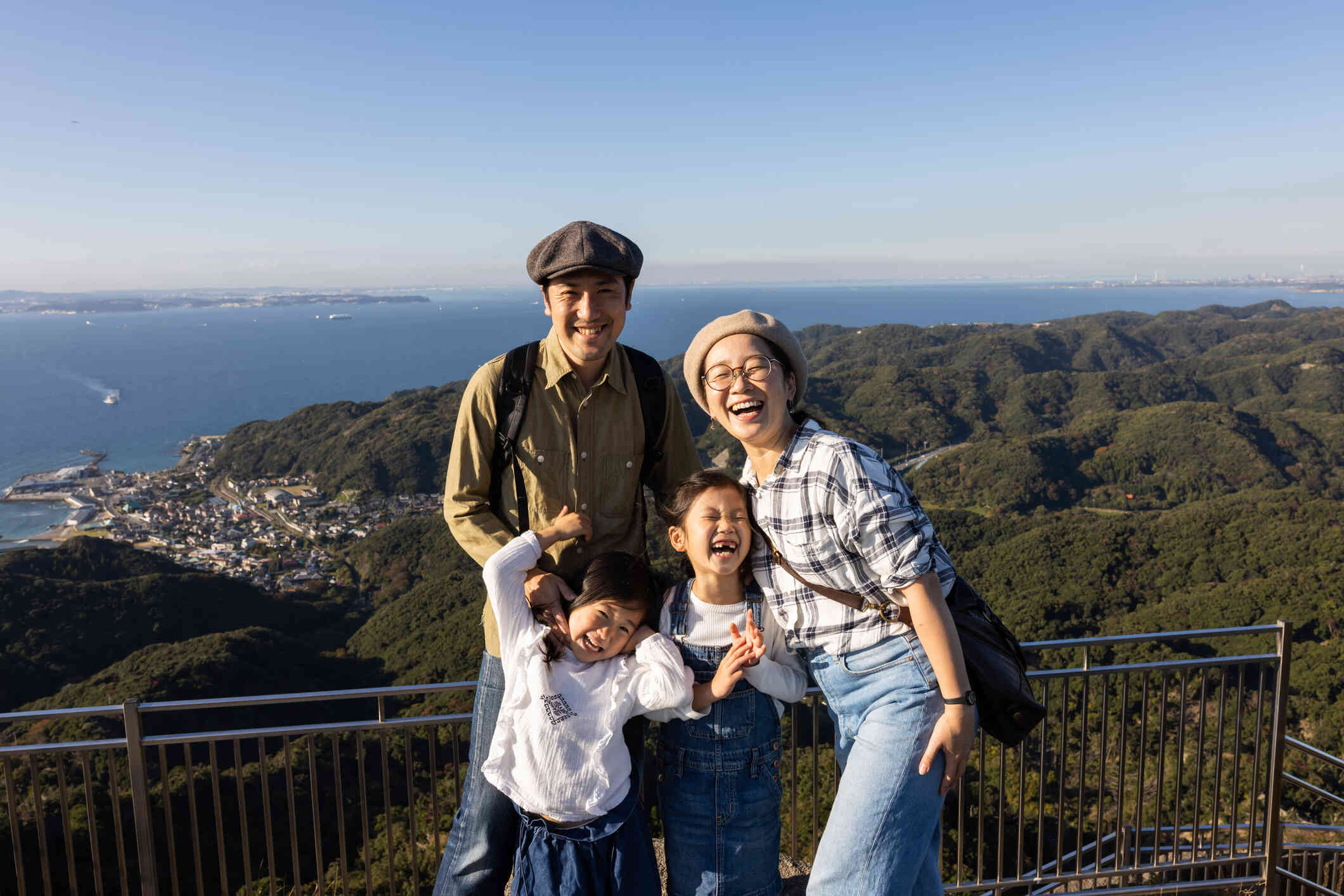 A mother, father, and two young children laugh while standing infront of a scenic view while looking at the camera.