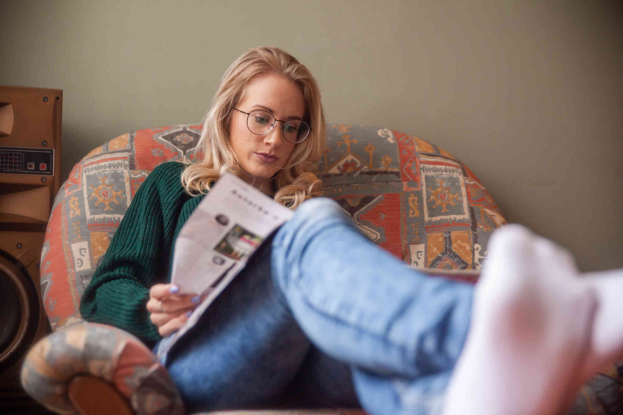 A woman with glasses sits in a large plush chair with her feet kicked up while looking at the magazine in her lap.