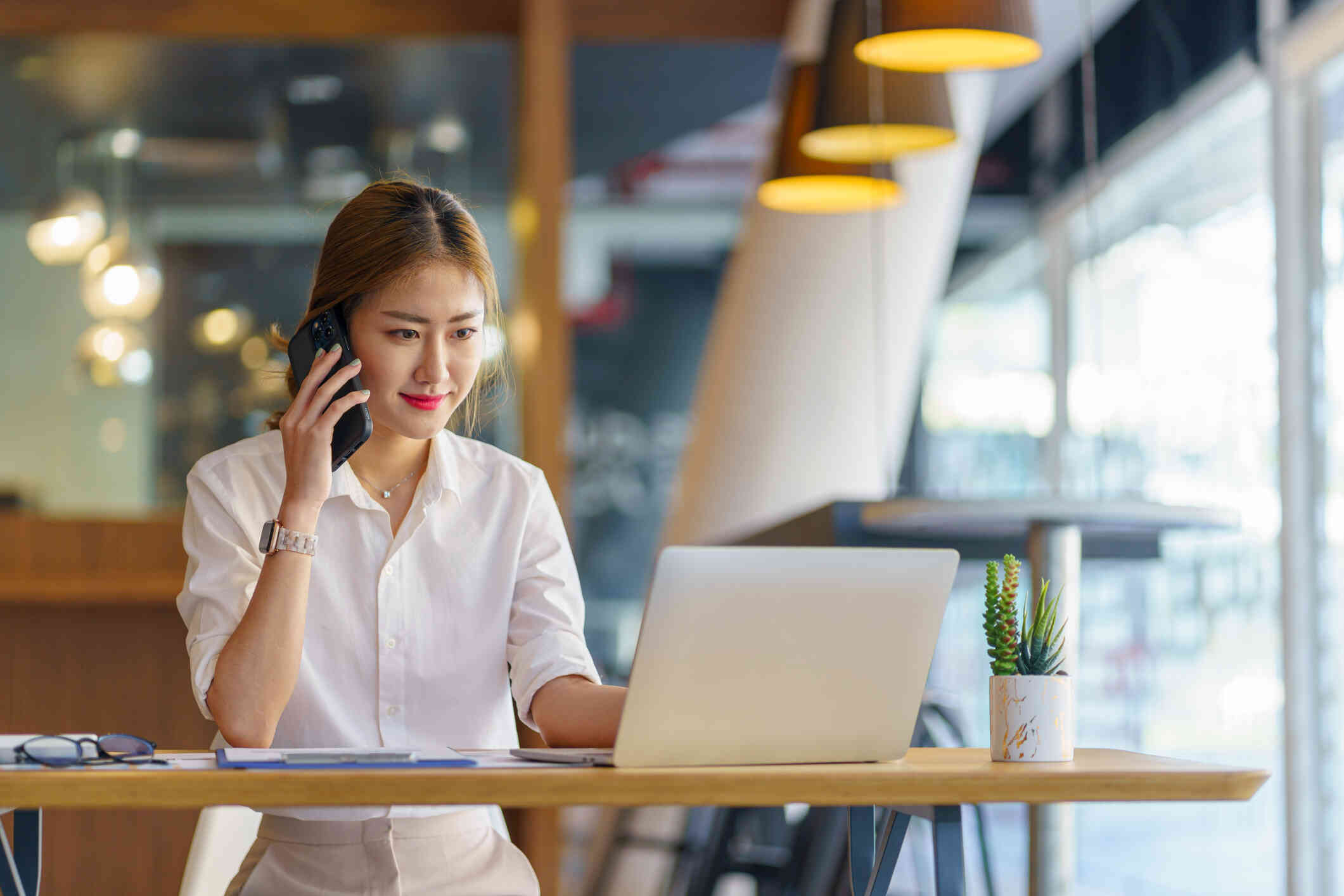 A woman in a white shirt sits at a table with her laptop open infront f her as she holds a phone to her ear.