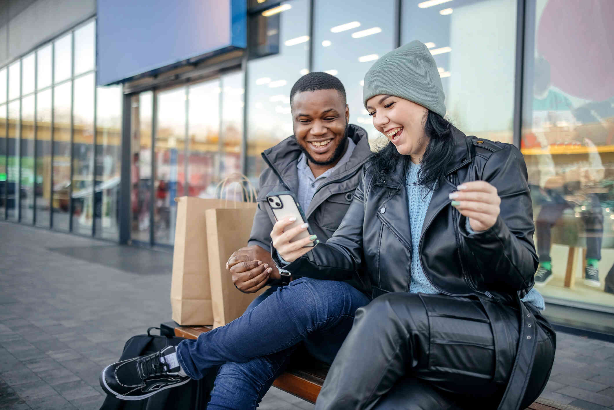 A male and female couple sit on a bech outside of a stire and look at a phone together while smiling.