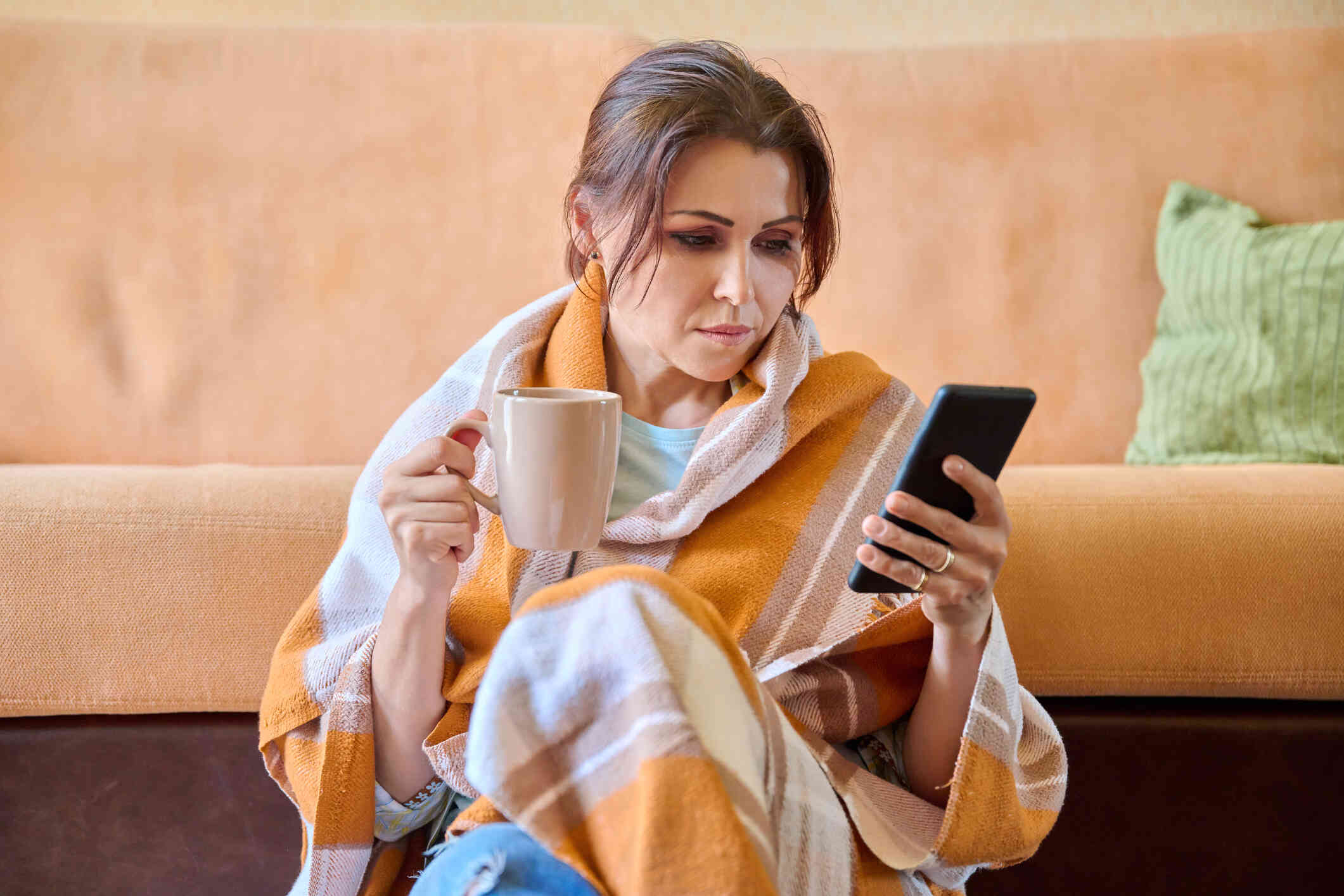 A woman sits on the floor with her back against a couch while holding a mug of coffee and looking at the cellhpone in her other hand with a serious expression.