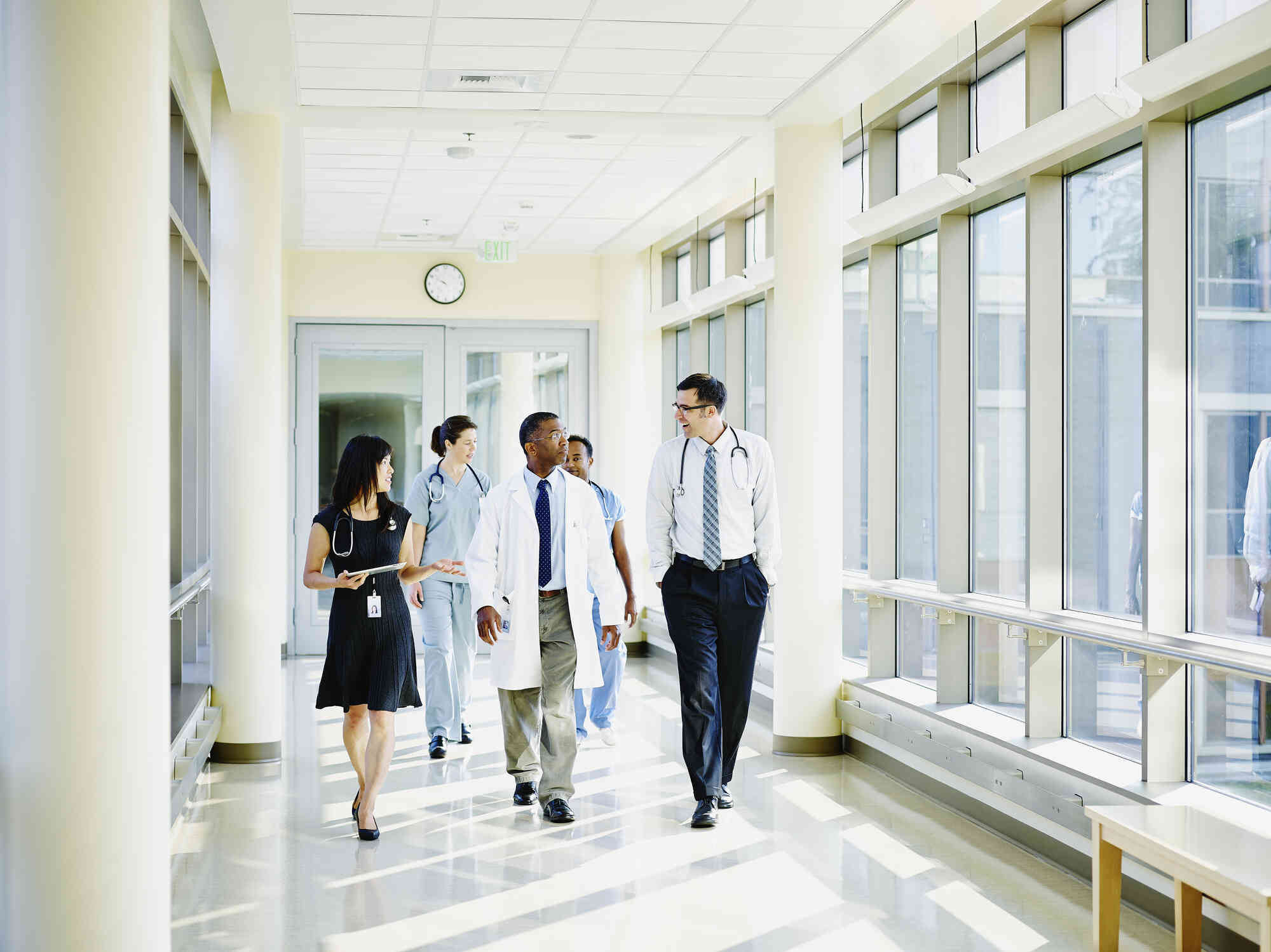 A man in a white coat walks down a hallway between a woman and a man with stethoscopes around their necks. Two nurses in scrubs walk behind them.