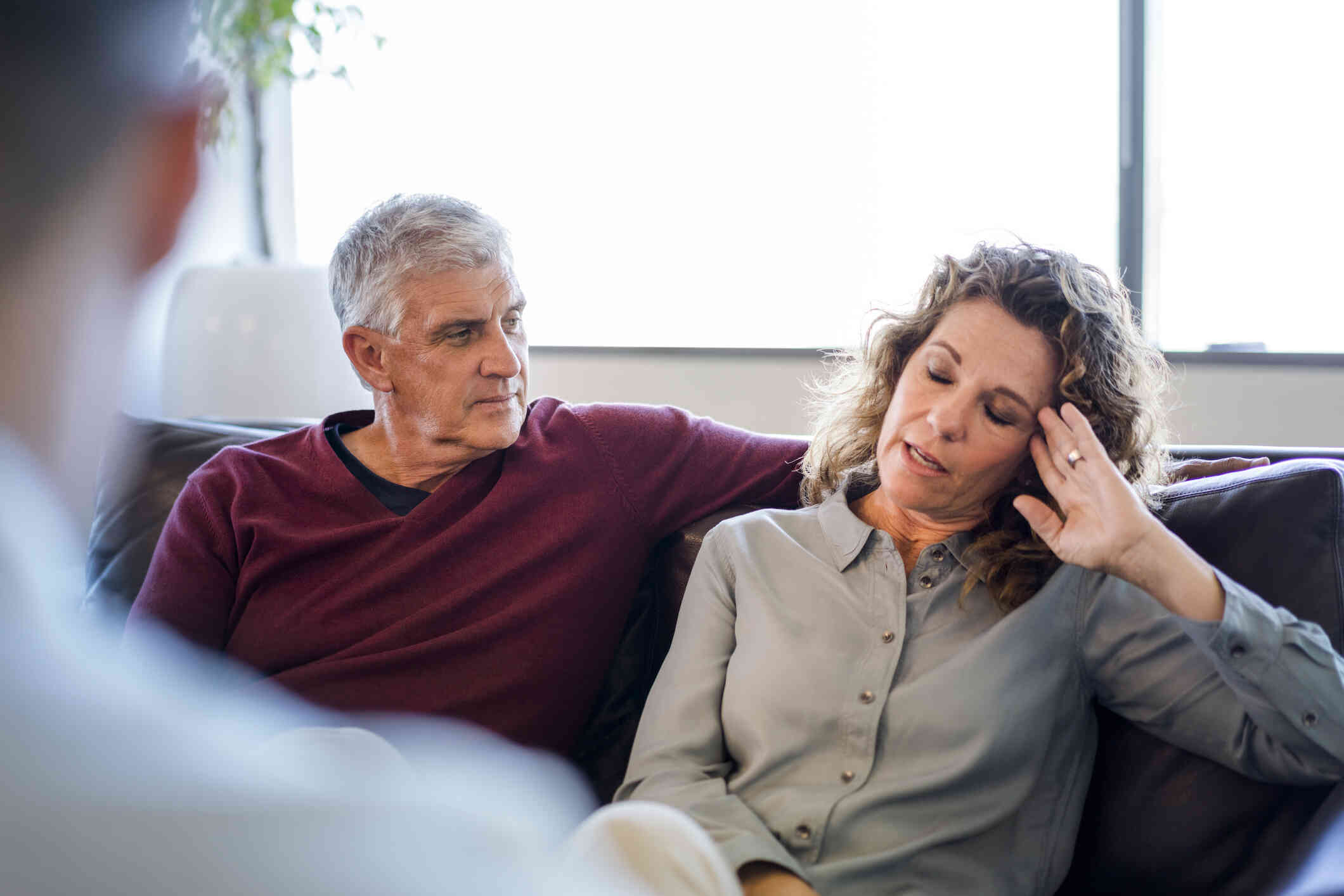 A man and a woman sit on a couch in an office and another person faces them with their back towards the camera. The woman closes her eyes and speaks with an upset expression.