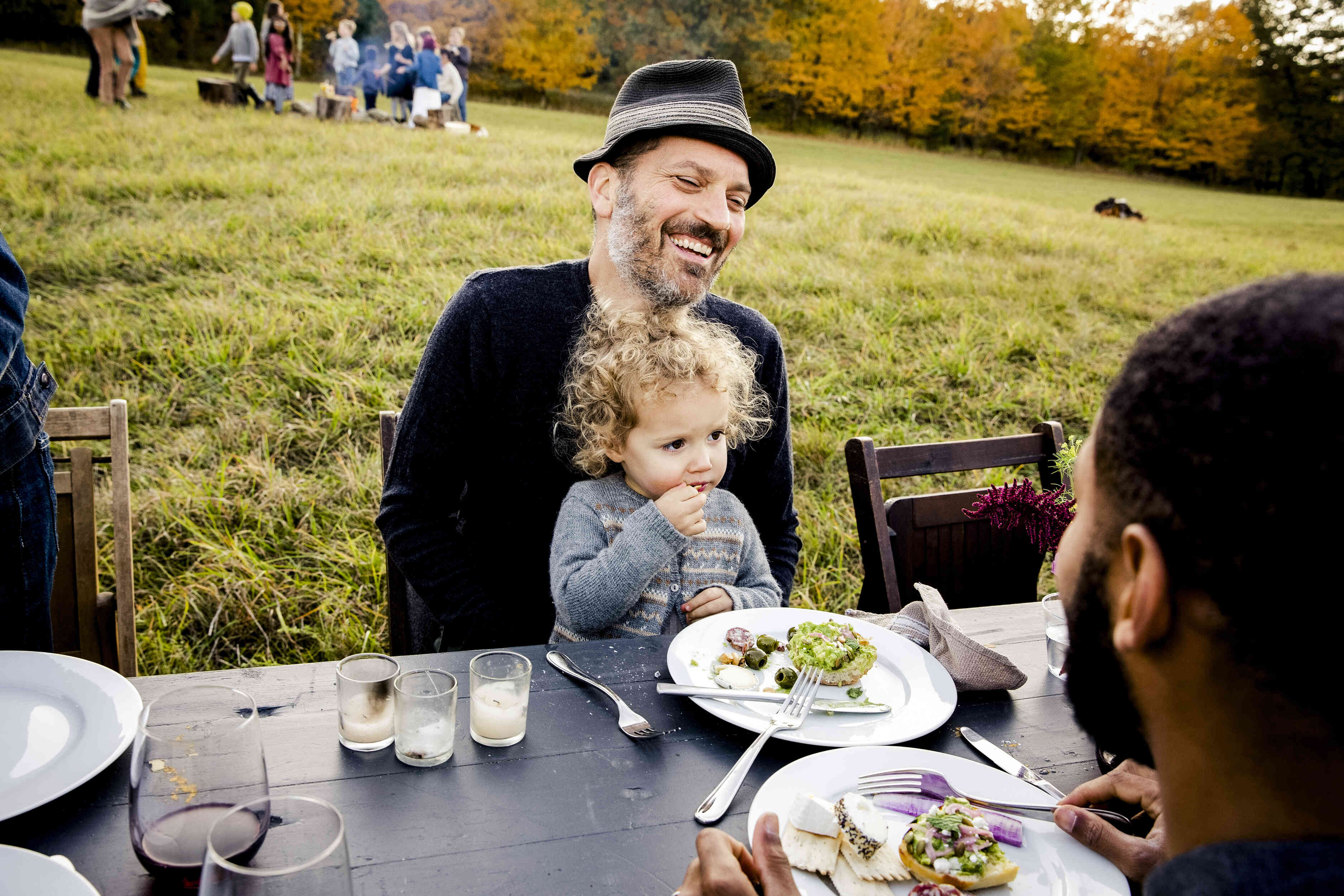 A man in a hat smiles as he sits at a table outside on the grass with plates of food in front of him and a blonde baby sitting on his lap.