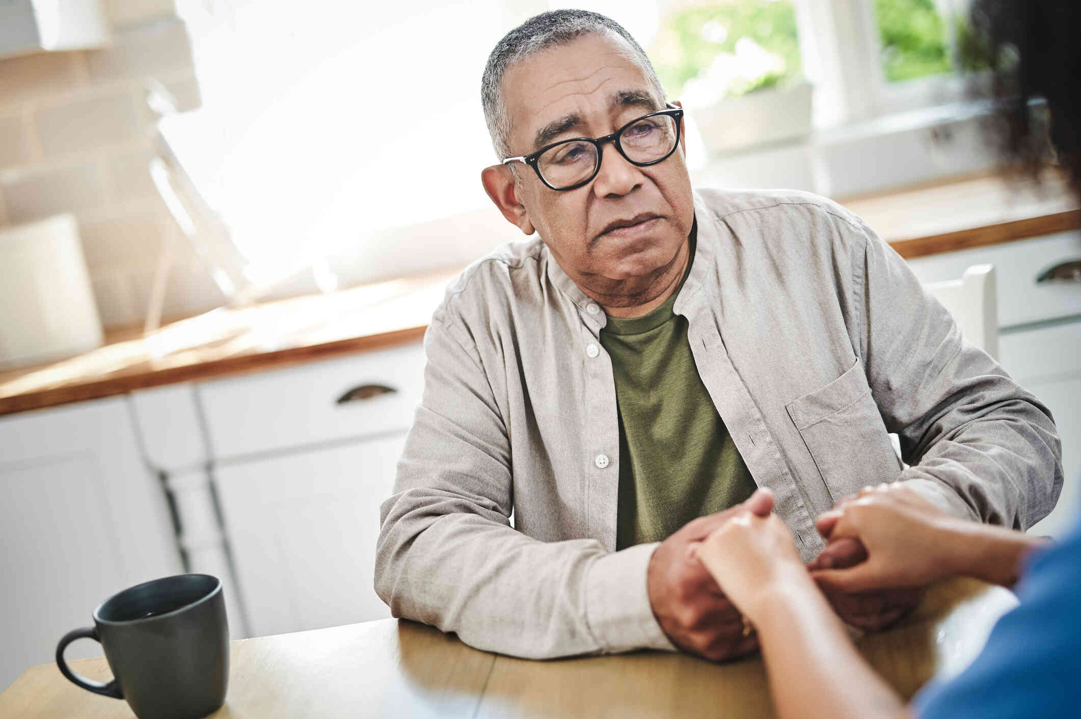 An elderly man with glasses sits at the ktichen table and holds the hands of the woman infront of him with a sad expression.