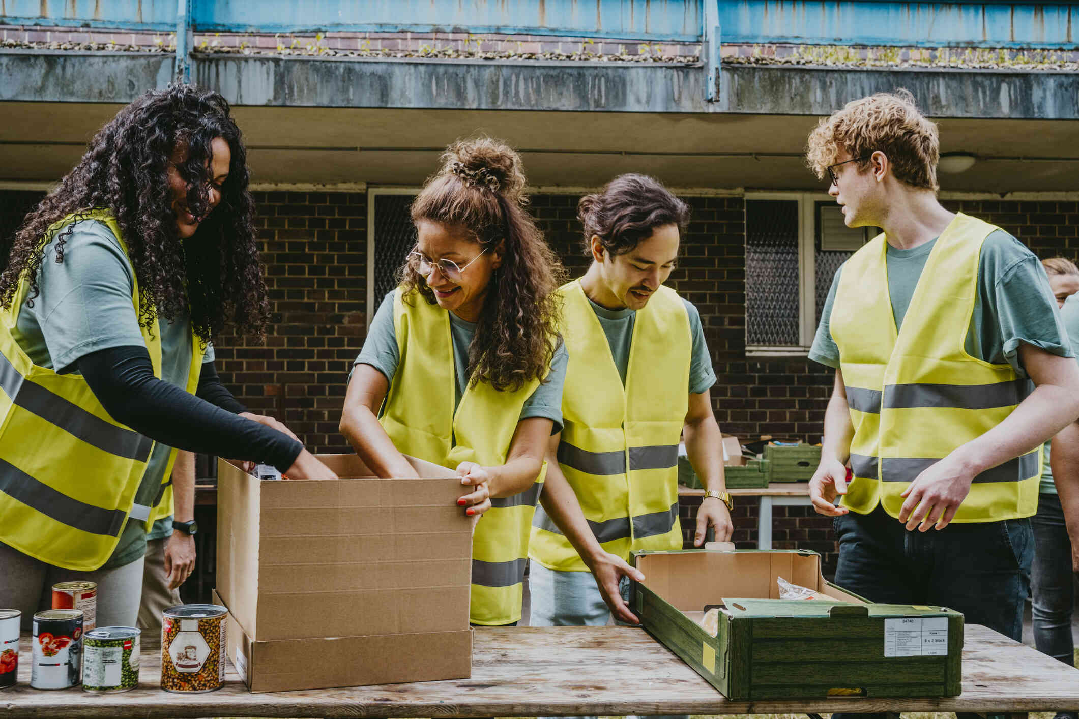 Four people in yellow safety vests smile as they stand outside and fill cardboard boxes with cans of food at a food bank.