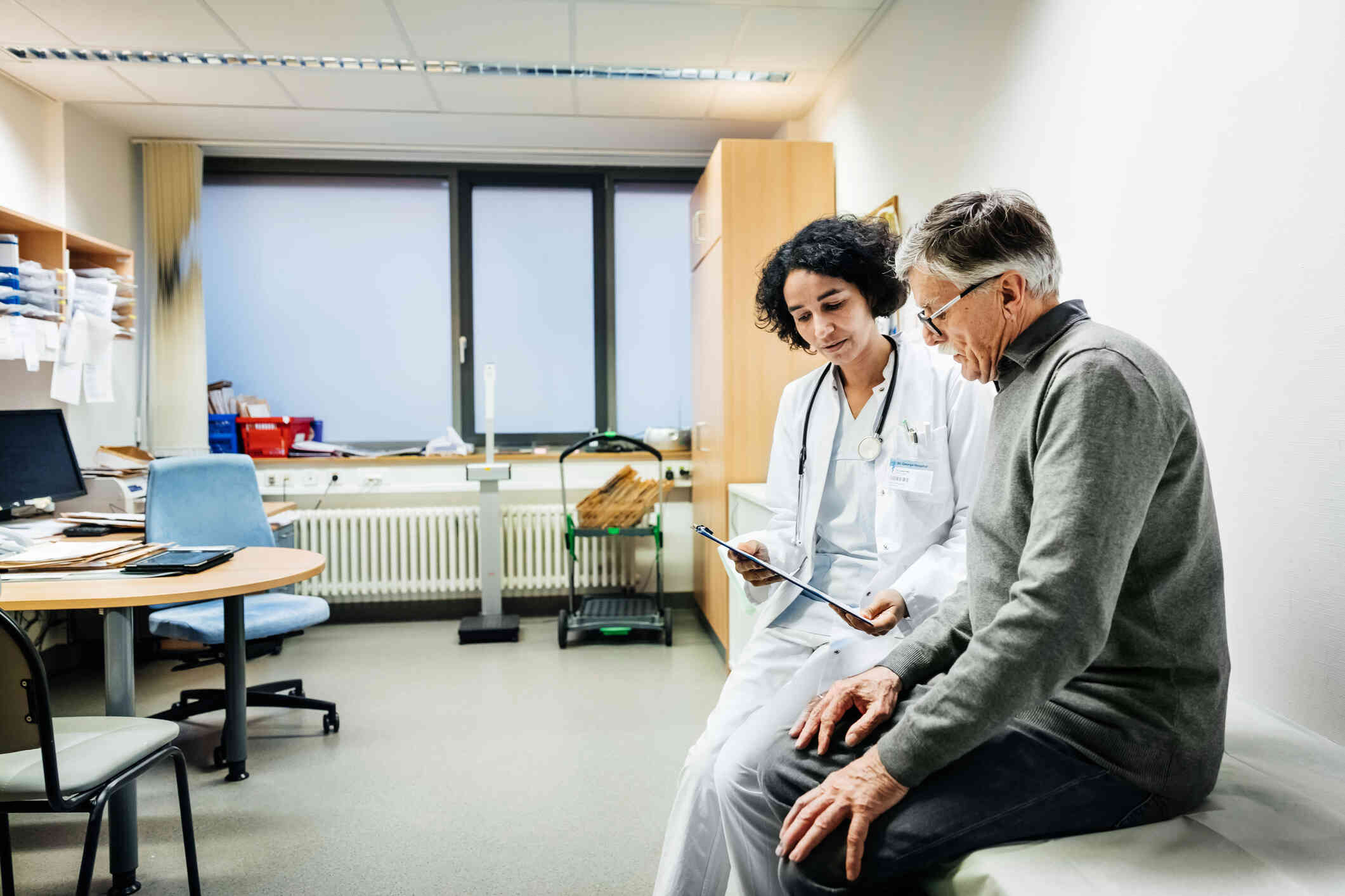 A female doctor sits next to her male patient on a medical table in the hospital while showing him some paperwork.