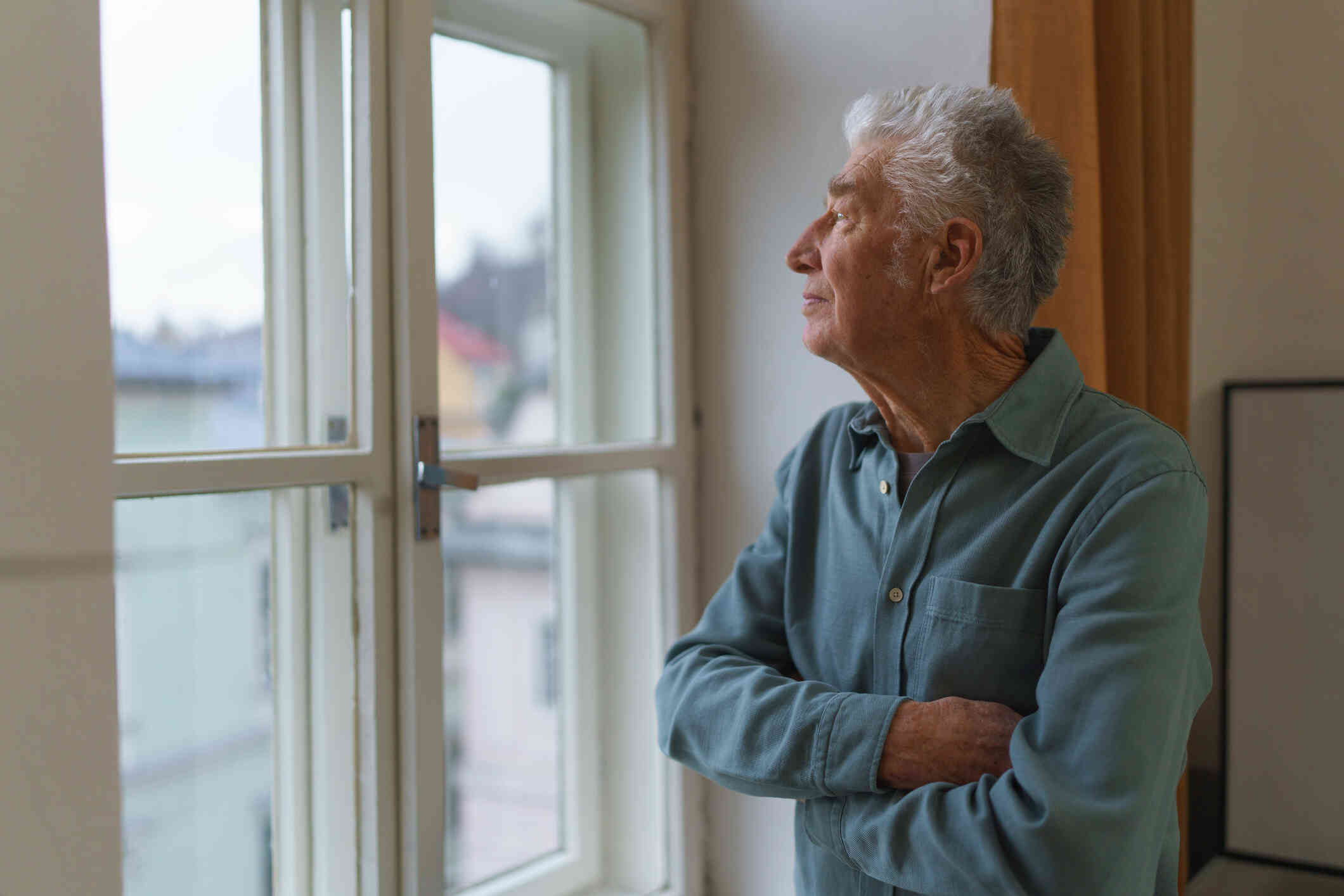 An elderly man in a button down shirt stands infront of a window of his home and crosses his arms across his chest while gazing out of the window sadly.