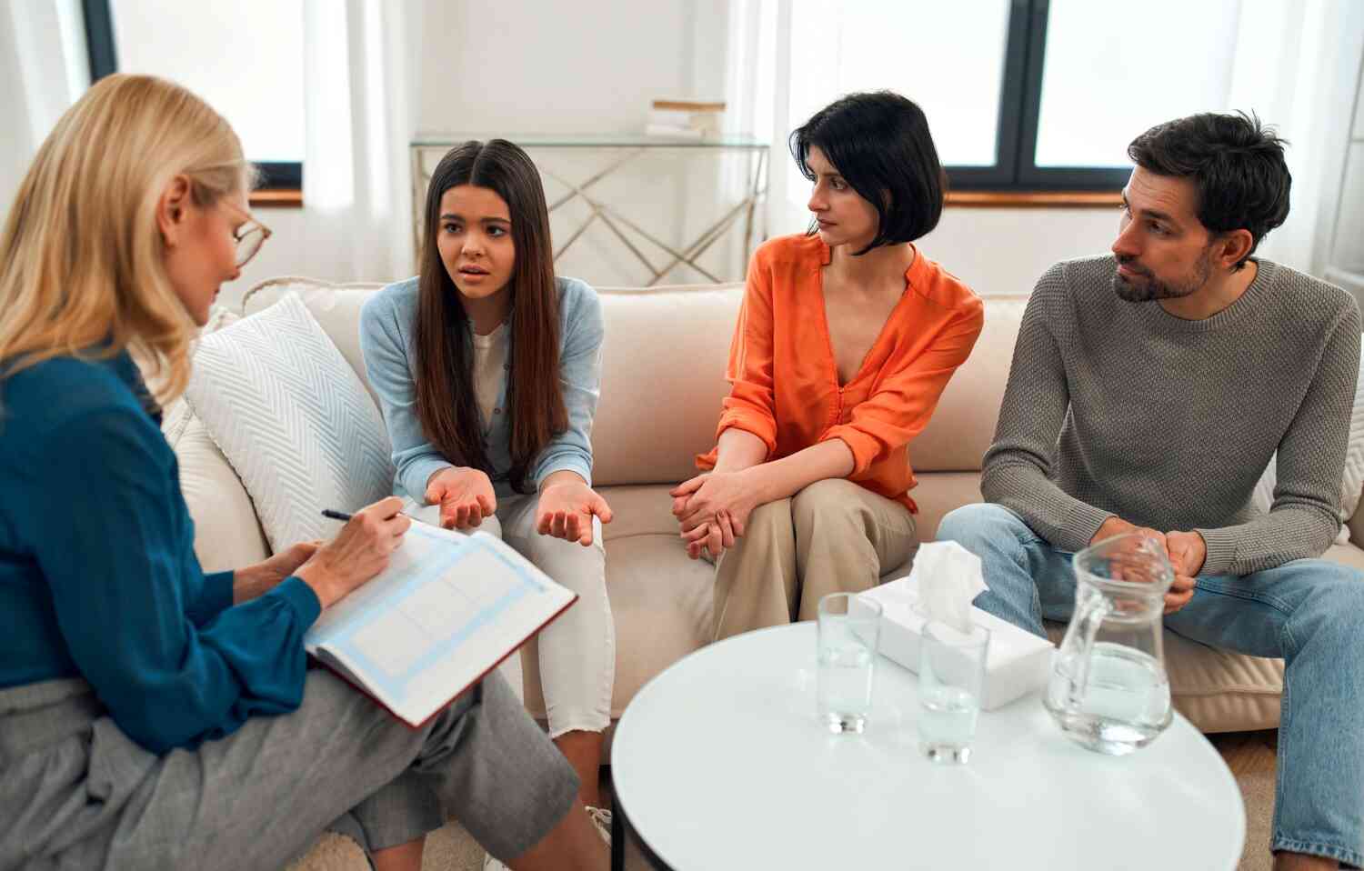 A teen girl in a blue shirt sits on the couch with her mother and father while talking with a therapist in a blue shirt taking notes