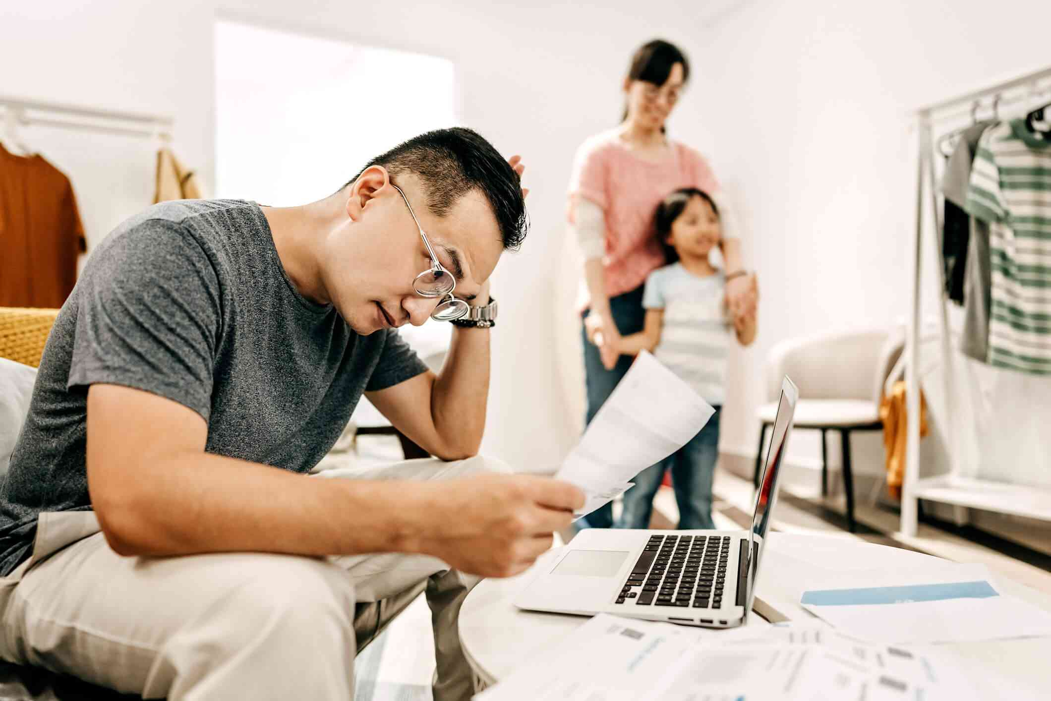 A man wearing glasses looks stressed as he sits on the couch with his laptop open infront of him and looks at some papers as his wife and child stand in the background.