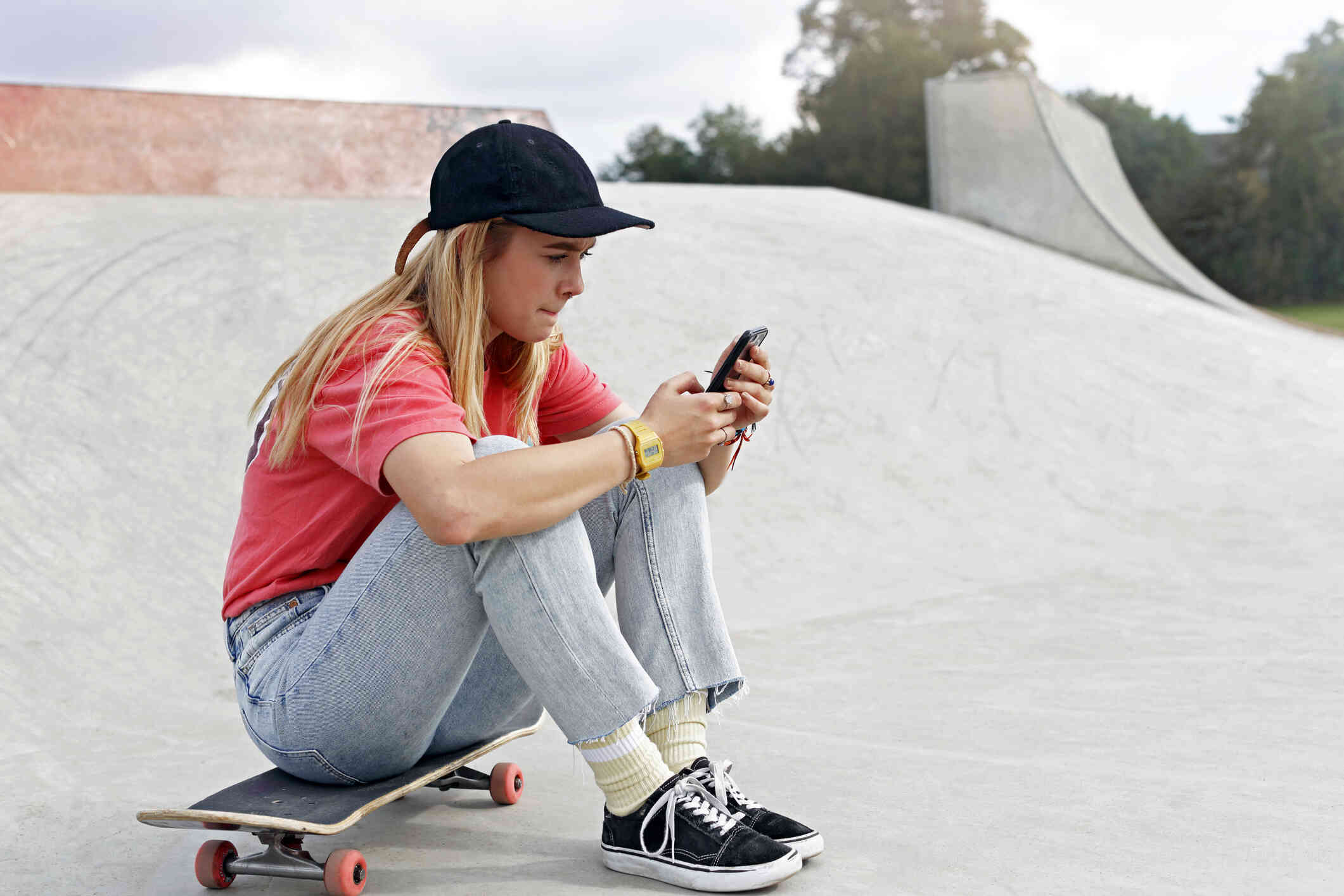 A teen girl in a baseball hat and red shirt sits on a skateboard and anxiously looks at a cellphone in her hand.
