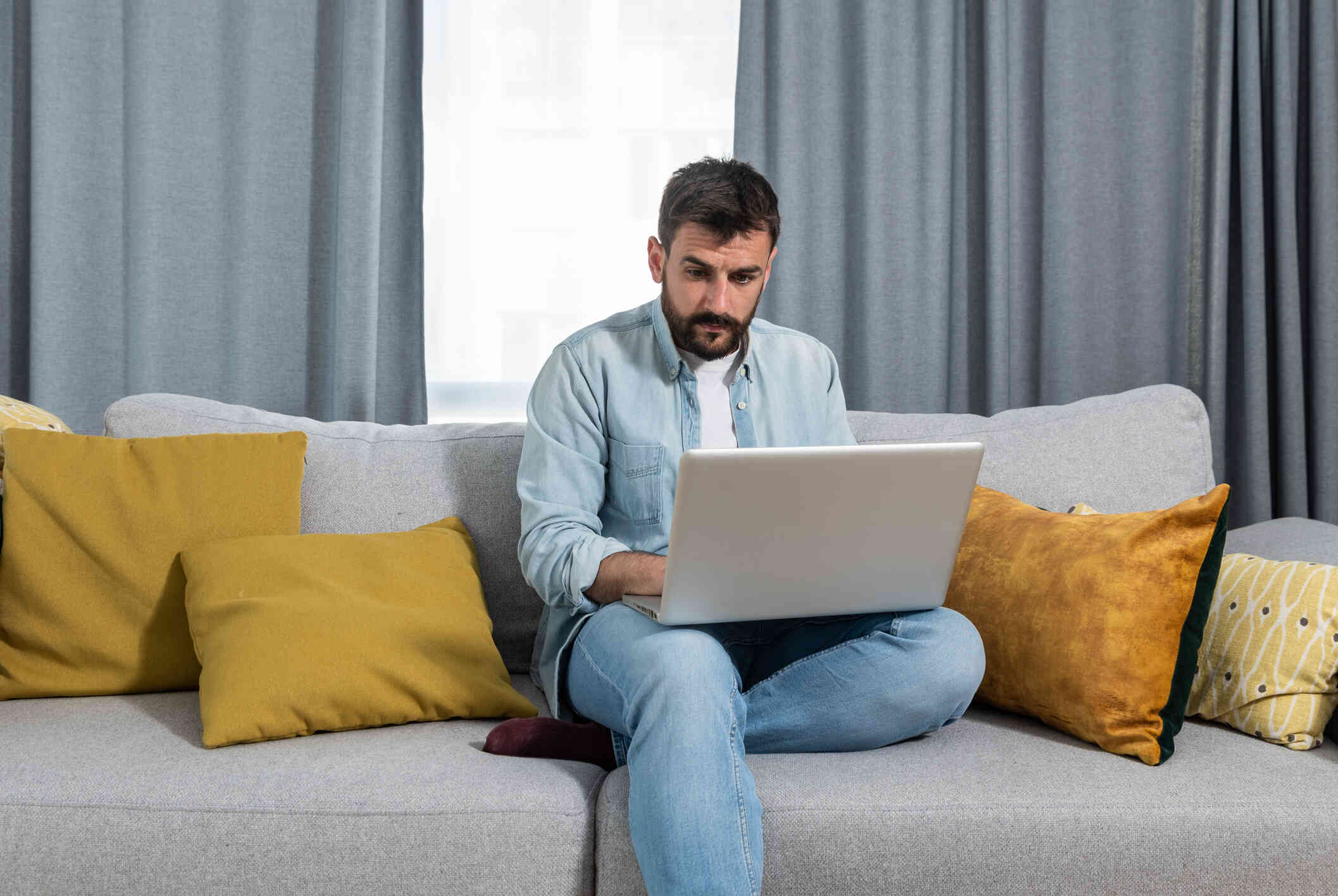 A man in a blue jean shirt sits on his couch and looks at  the laptop open in his lap with a serious expression.