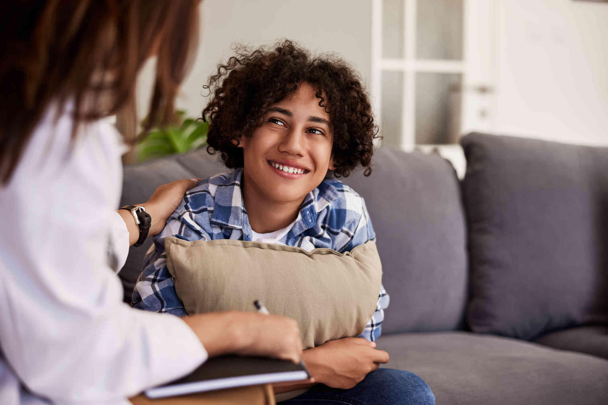 A young boy sits on a couch and clutches a pillow to his chest while smiling up at the therapist sitting across from him.