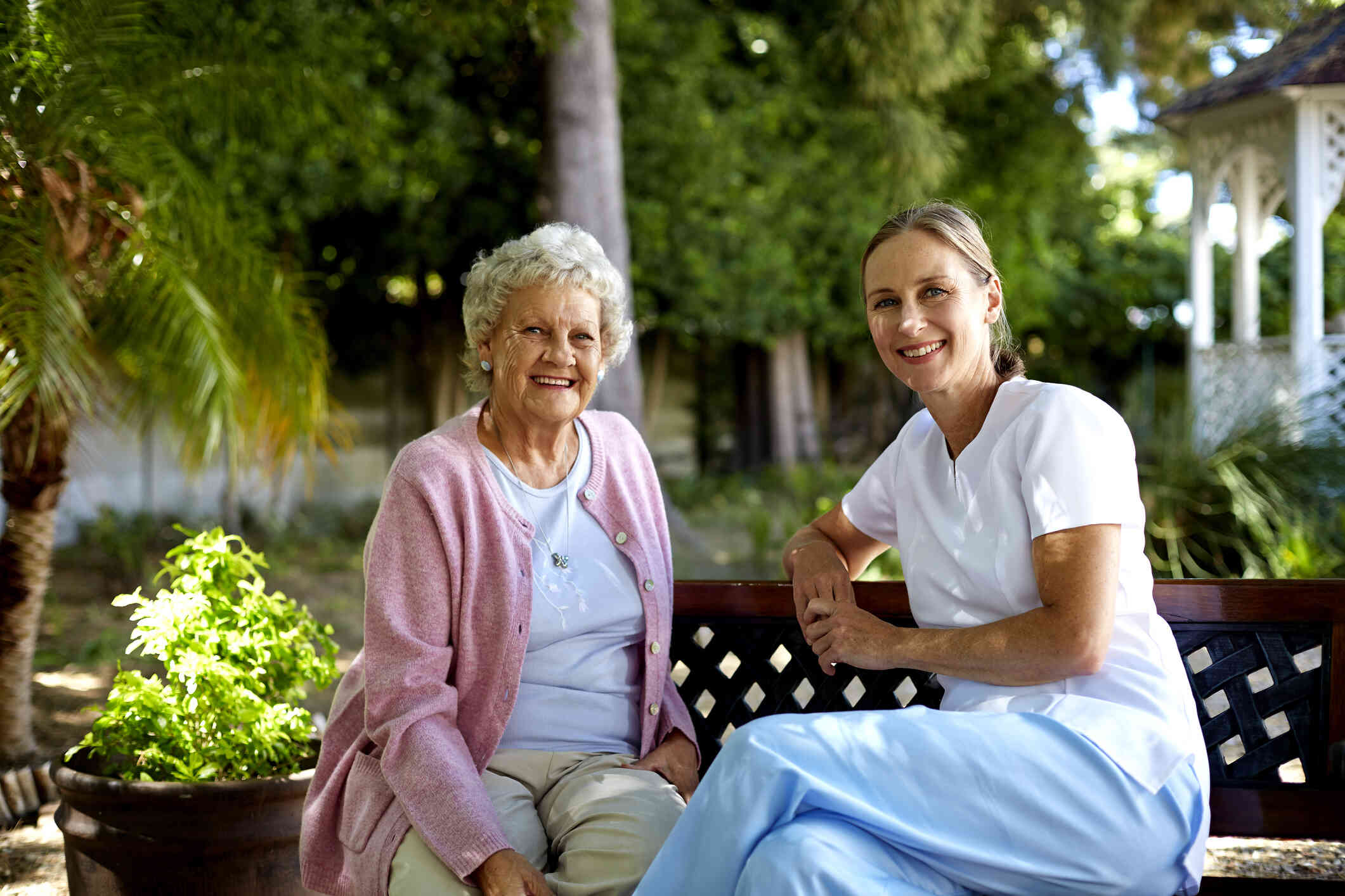 An elderly woman sits on a park bench on a sunny day  next to a younger woman in scrobs as they both smile at the camera.