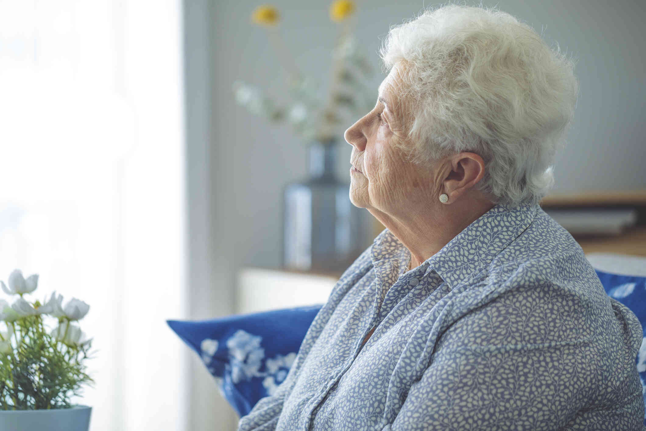 A close up of an elderly woman as she sits in her home and gazes off.