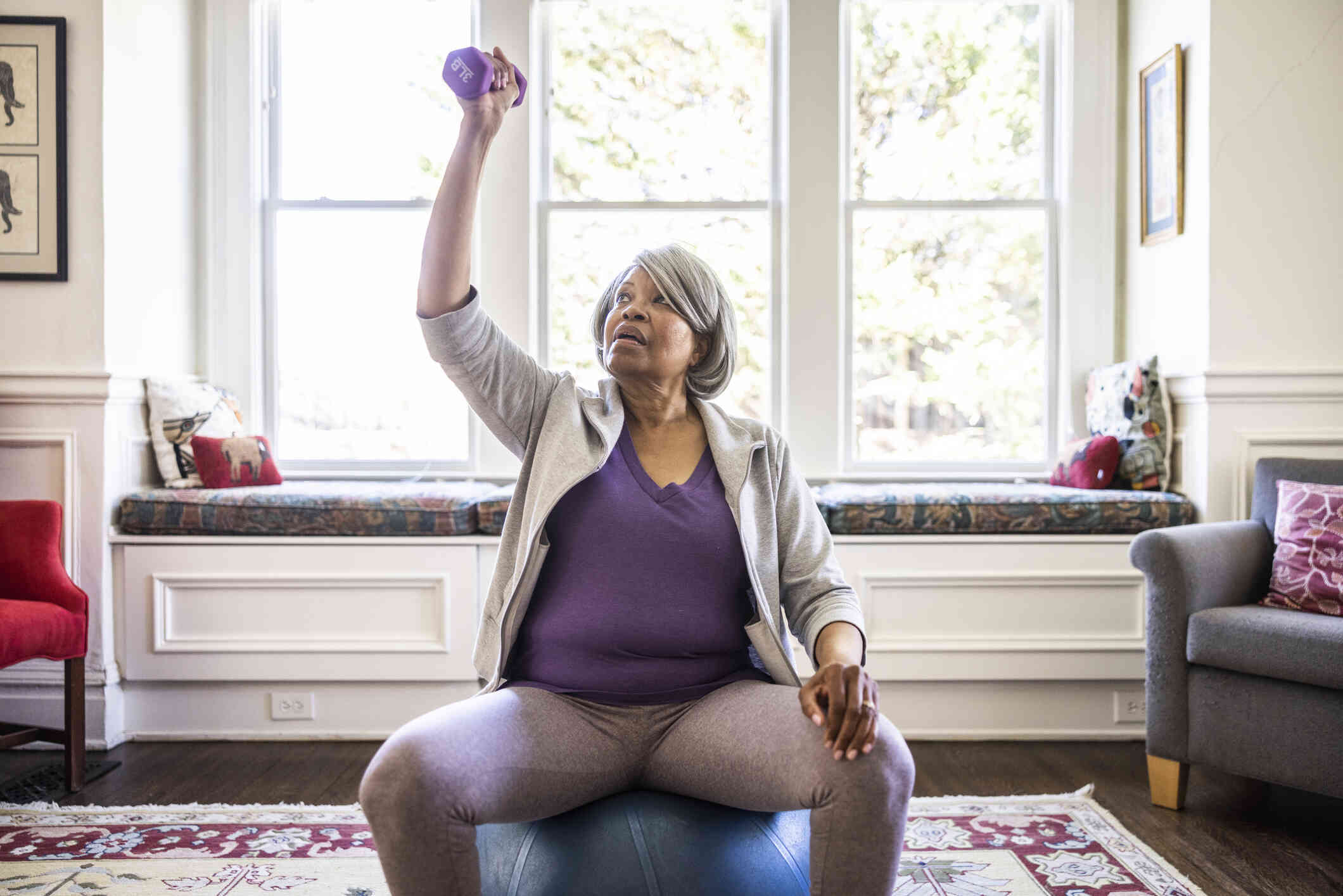 An elderly woman in a purple shirt sits on a workout ball in her home while lifting purple 3 pound weights.