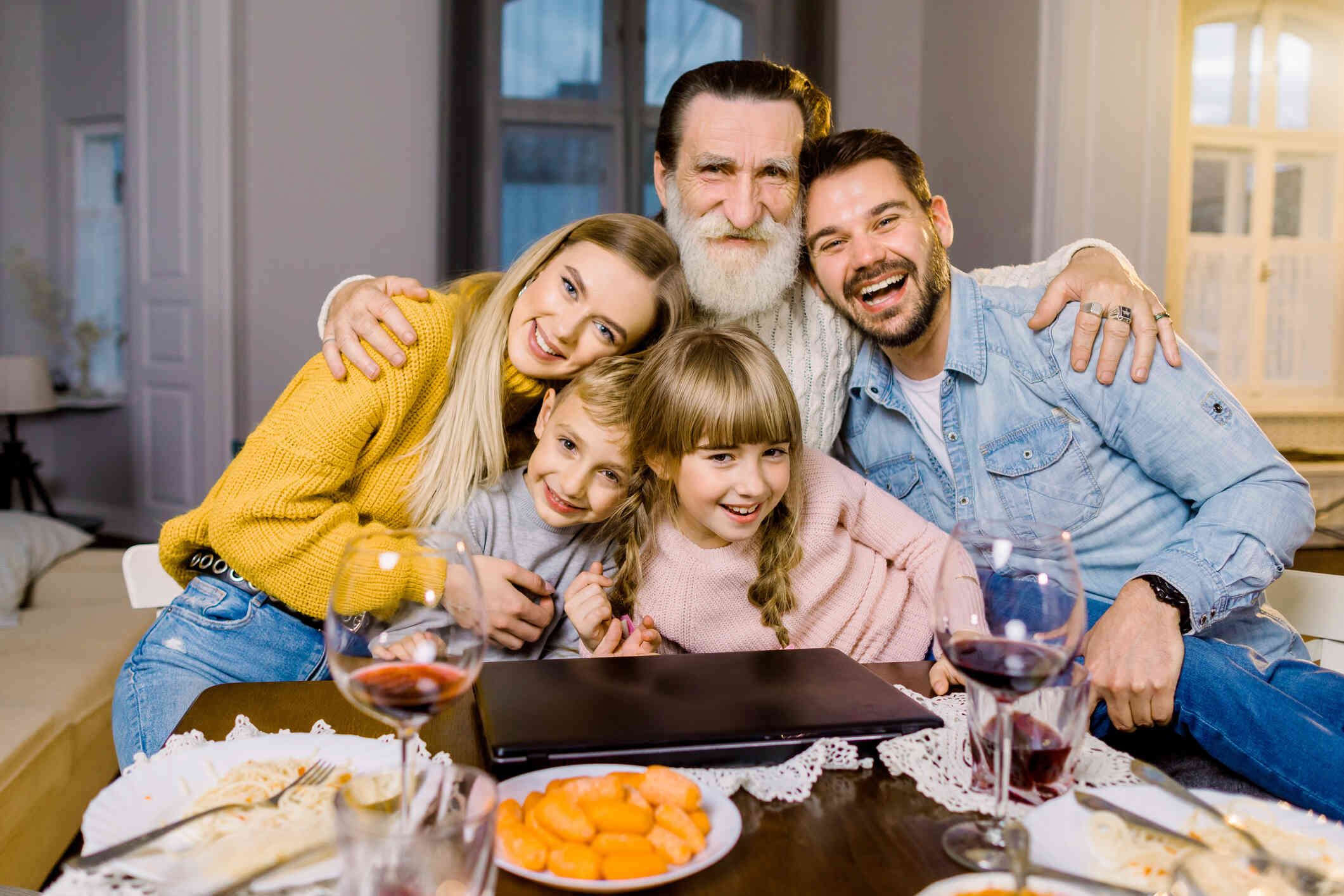 A mature man with a gray beard smiles and hugs two adults and two children as they sit in front of a table with two glasses of red wine on it.