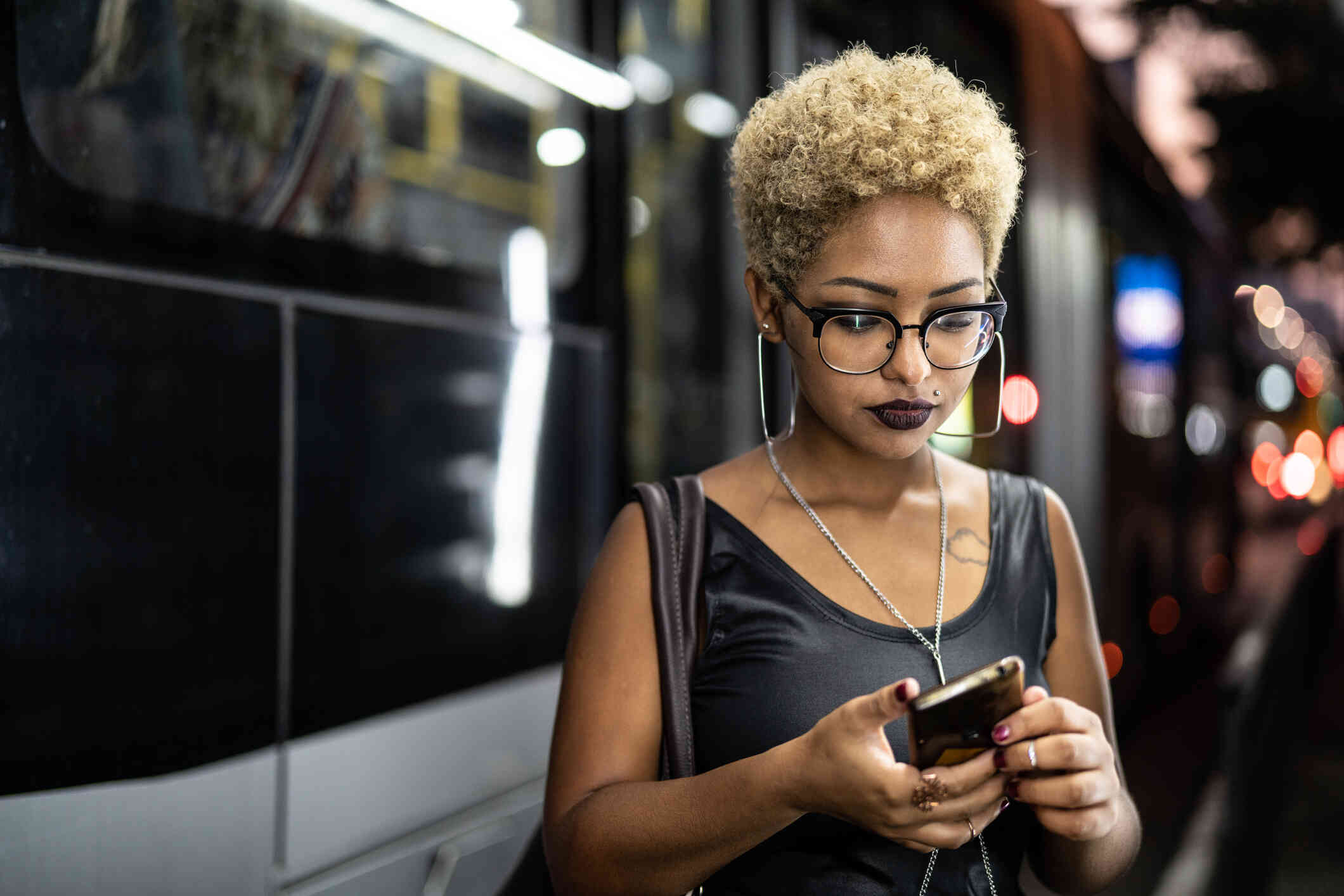 A woman with glasses in a black shirt stands in the subway and looks at the cellphone in her hand with a serious expression.
