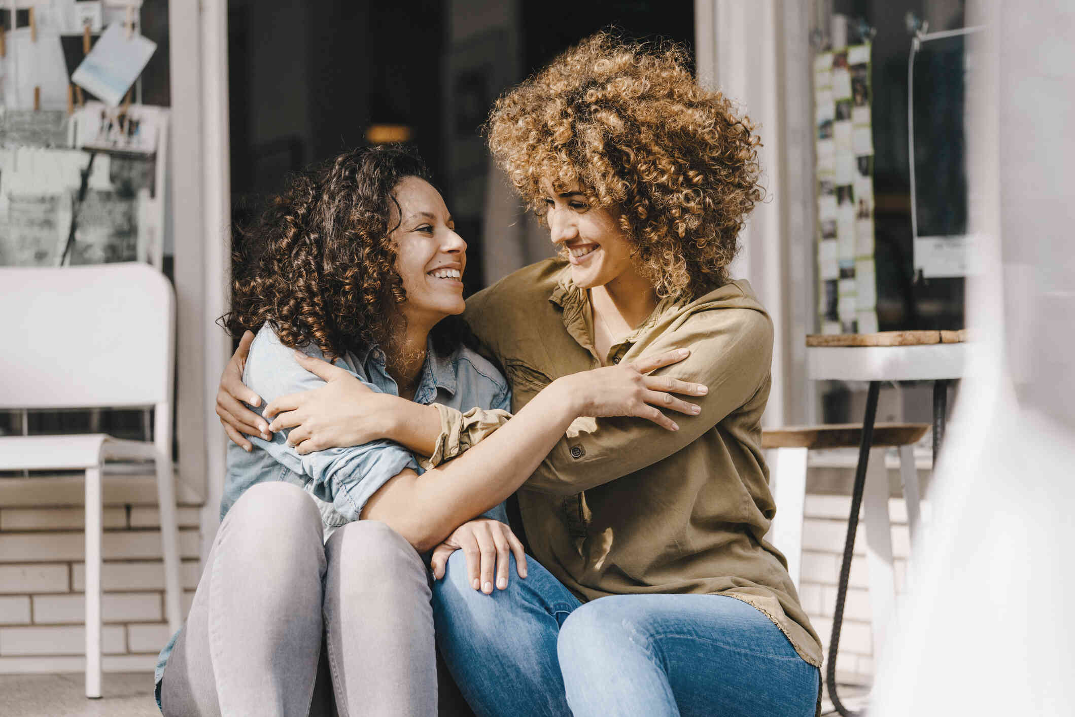 Two female adult family members wrap their arms around each other while smiling at each other as they sit outside on porch steps on a sunny day.