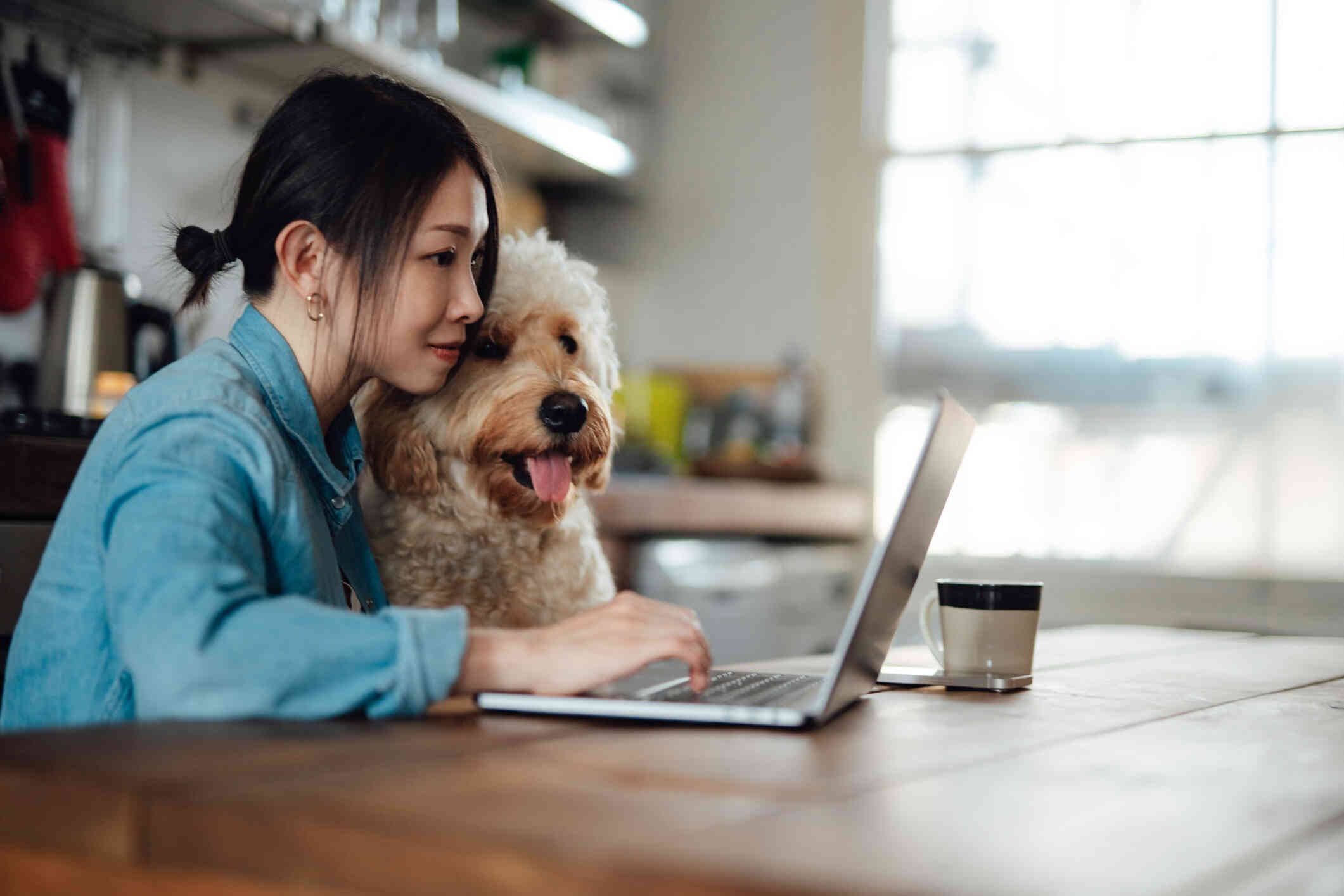 A woman sits at the kitchen table with her dog next to her as she looks at the lapto open on the talbe infront of her.