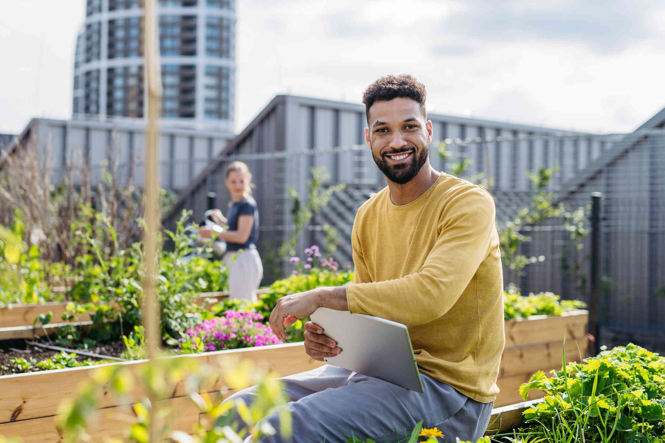 A man in a yellow shirt sits outside with his laptop on a rooftop surrounded by a garden as he smiles at the camera.