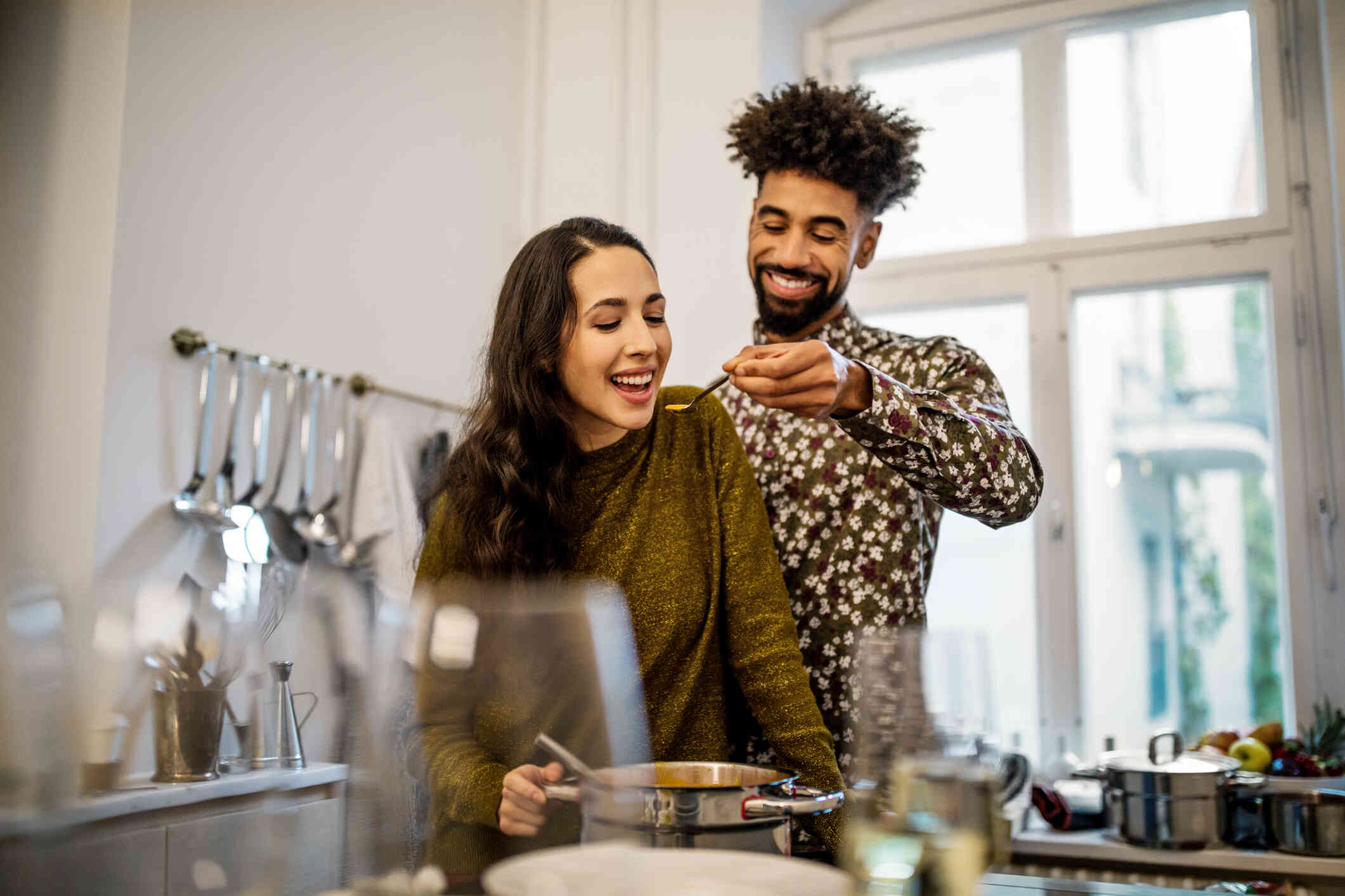 A man smiles as he stands in a kitchen and feeds a woman standing next to him a taste of food on a spoon.